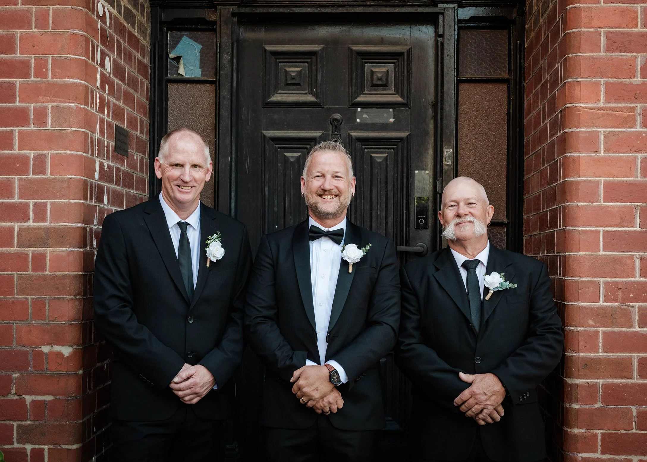 Groom in a black tuxedo posing with two groomsmen in black suits in front of a brick building entrance
