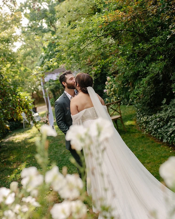 A bride and groom sharing a kiss in a lush, green garden with trees, bushes, and flowers, during daytime.