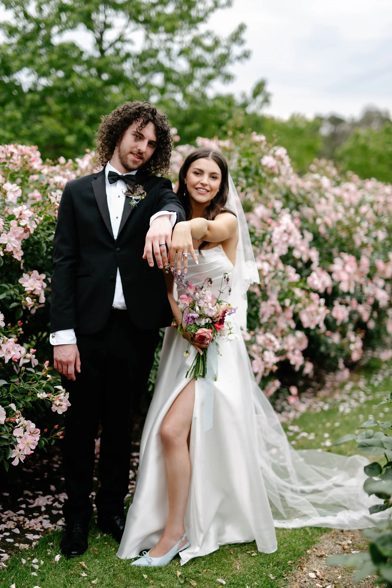 Bride and groom posing outdoors among pink flowering bushes, showing wedding rings, with the bride holding a bouquet and the groom in a black tuxedo with a bow tie.