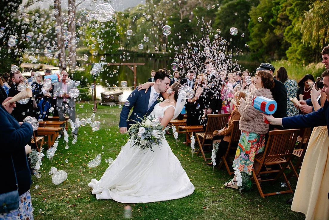 A newlywed couple kisses during their outdoor wedding celebration while guests throw bubbles and take pictures, surrounded by trees and chairs.