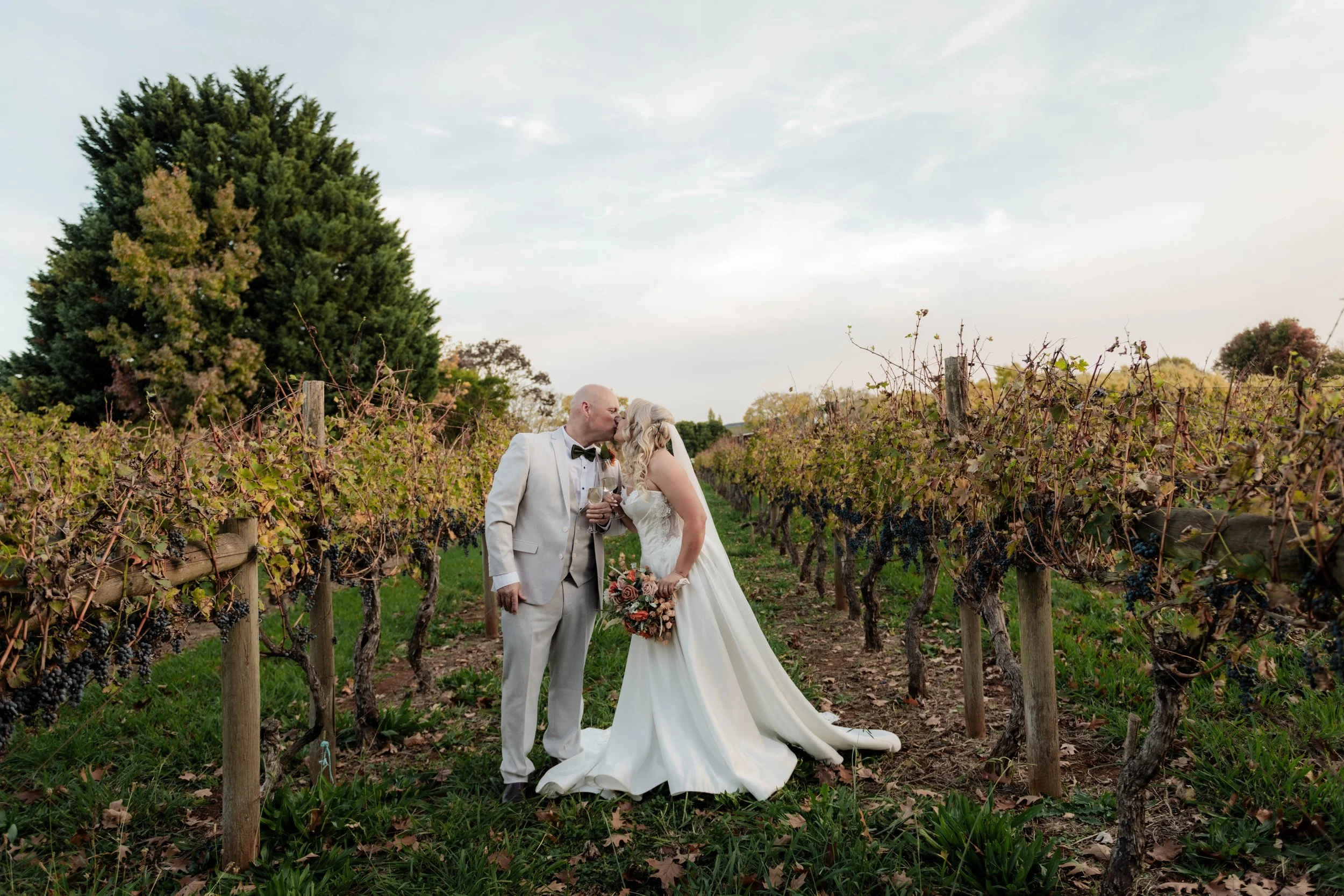 A newlywed couple shares a kiss in a vineyard, with the groom in a white tuxedo and the bride in a white wedding dress holding a bouquet, surrounded by grapevines under a sky with scattered clouds.