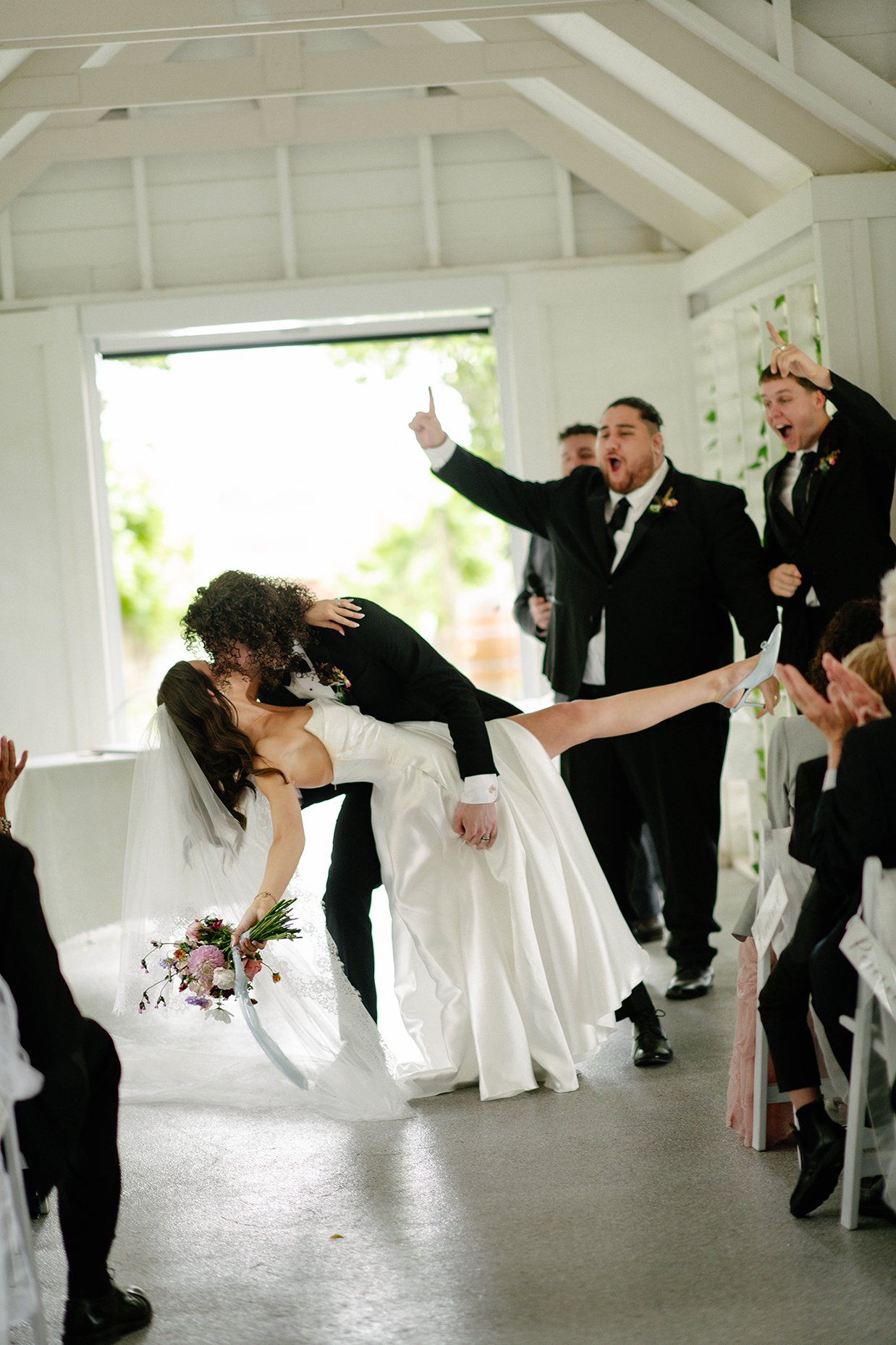 Groom dipping bride during wedding in a bright, white venue with guests applauding.
