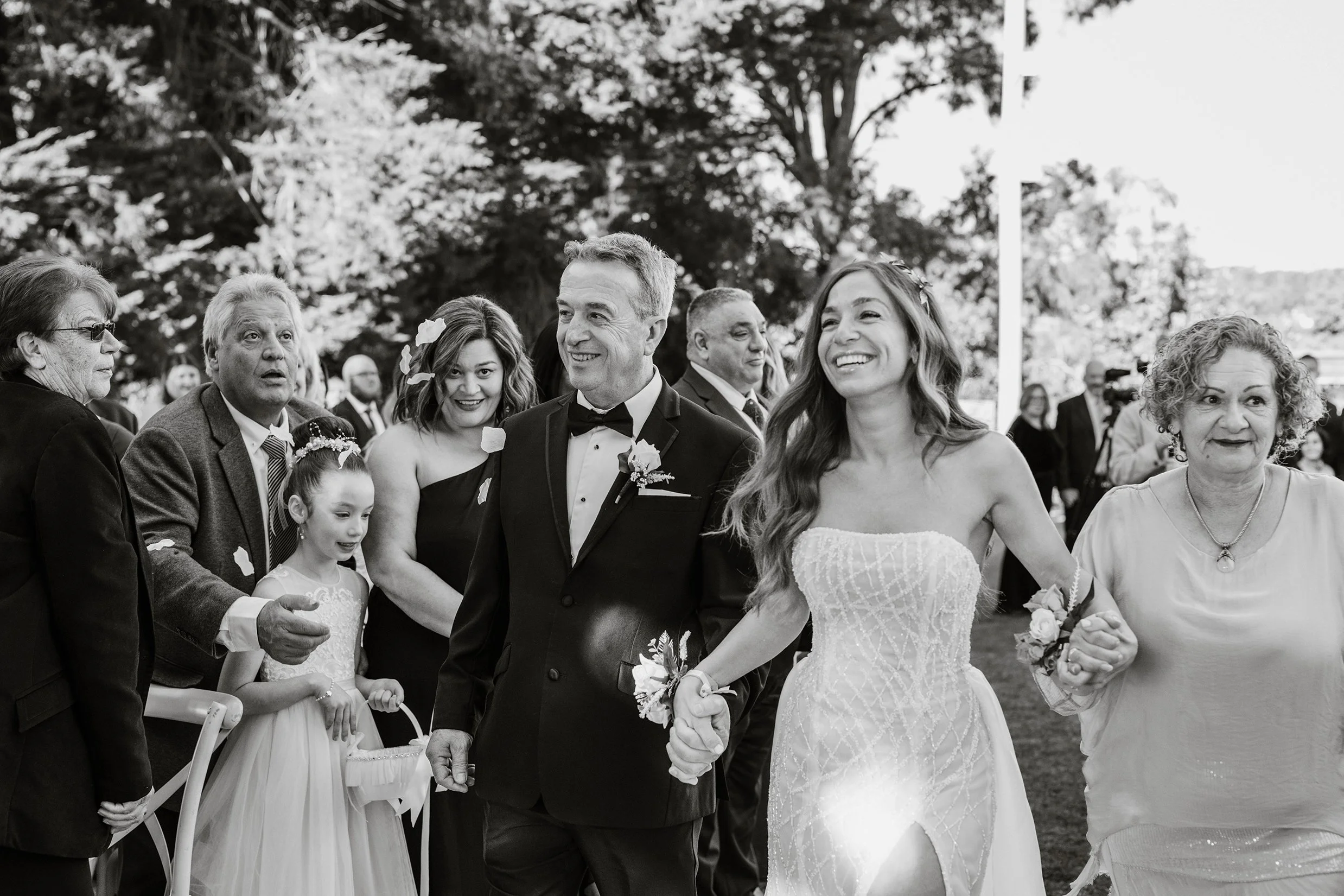 Black and white photograph of a wedding celebration outdoors with a bride and groom walking, holding hands. The bride is smiling, wearing a strapless wedding dress, and the groom is wearing a tuxedo with a bow tie. They are surrounded by guests, incl