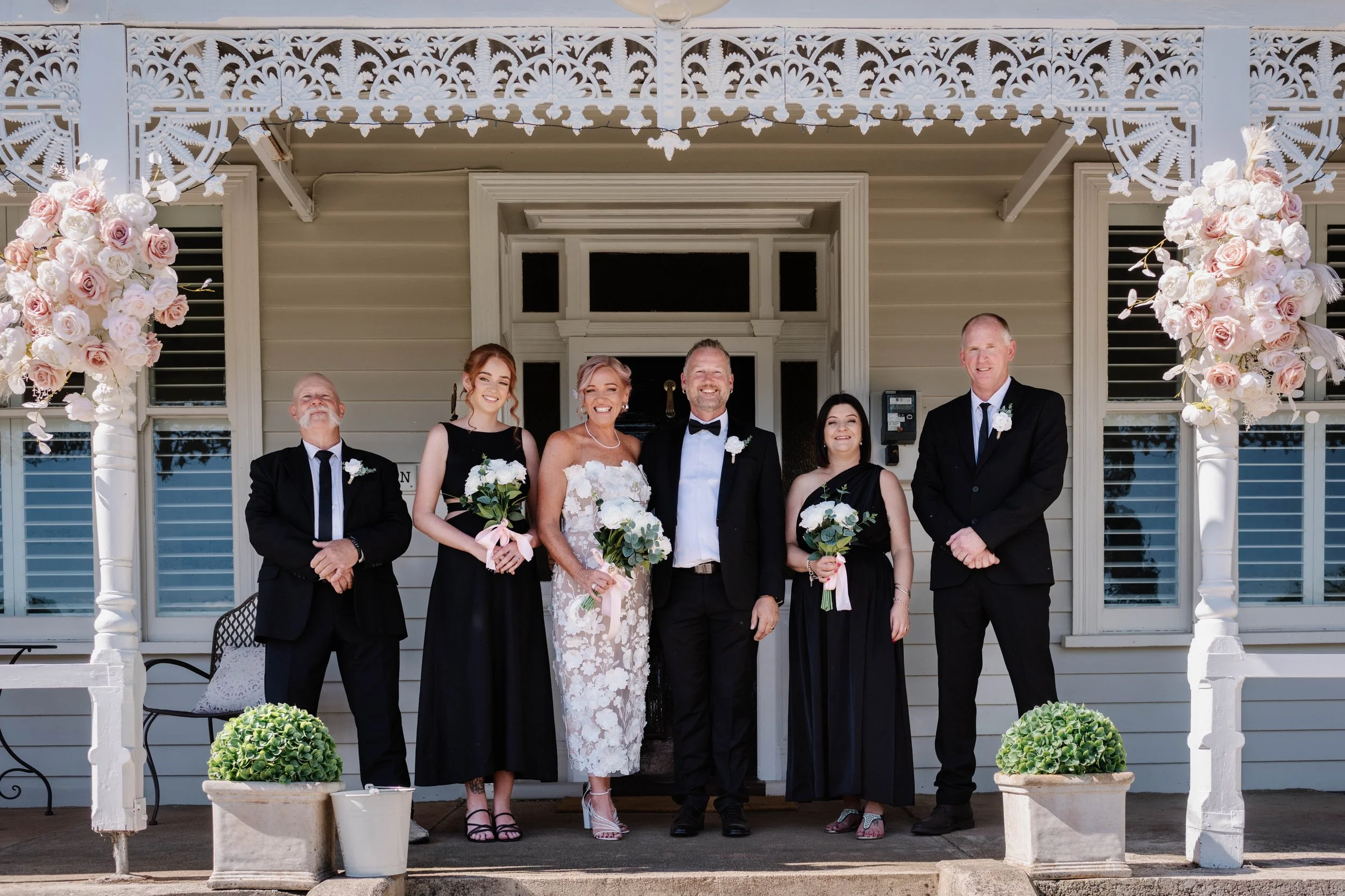 Bride and groom posing with wedding party members on the front porch of a historic-style wedding venue framed by floral arrangements
