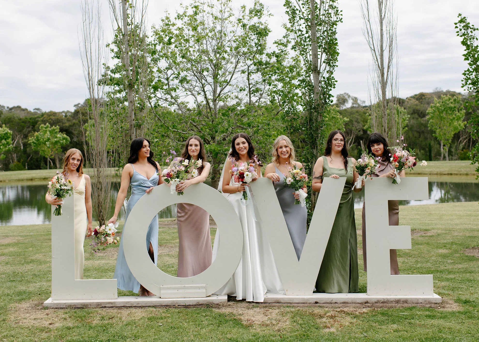 Six women in formal dresses holding bouquets of flowers standing behind large white letters spelling out 'LOVE' by a pond in a park with trees and a cloudy sky