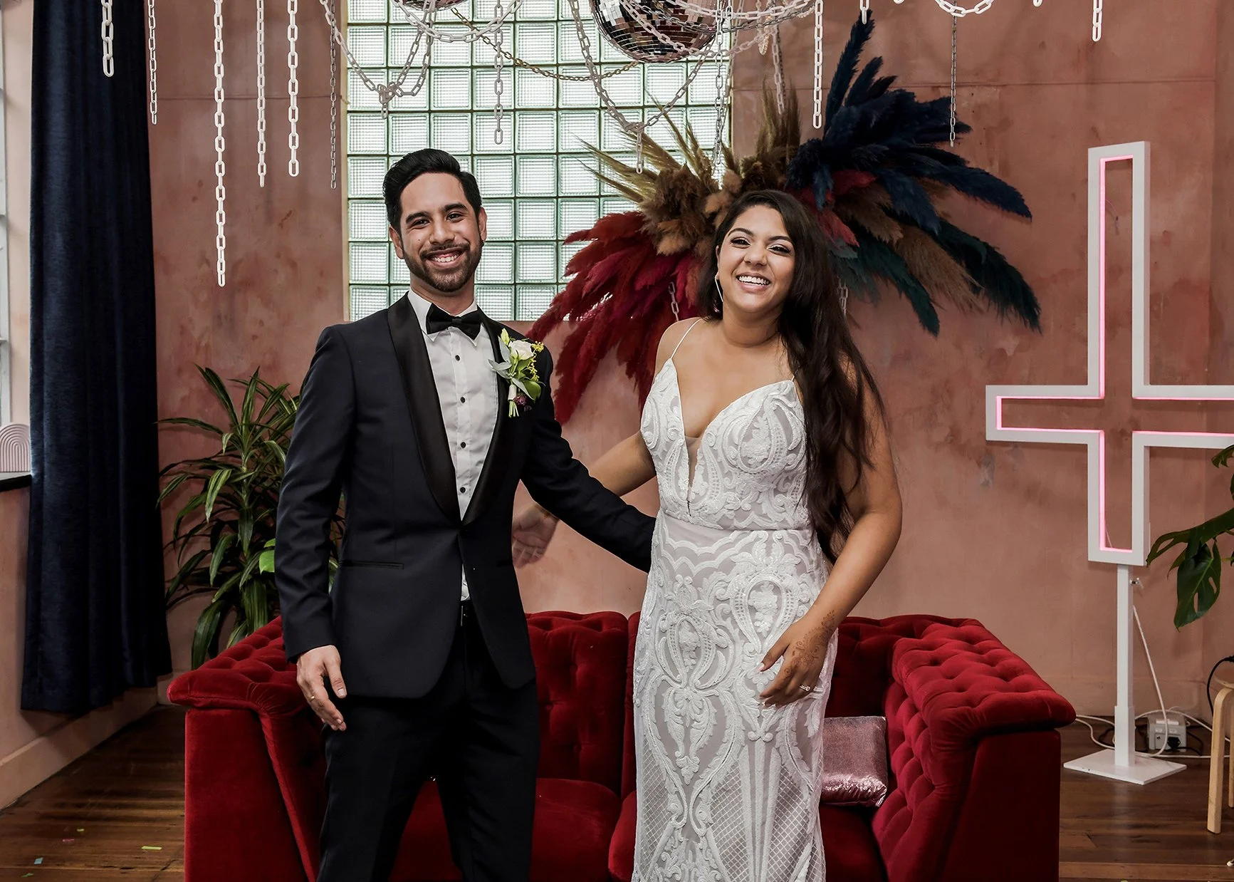 A newly married couple standing in front of a decorative wall, smiling at the camera. The groom wears a black tuxedo with a bow tie, while the bride wears a white lace wedding dress. Behind them is a colorful feathered headdress and a pink cross-shap