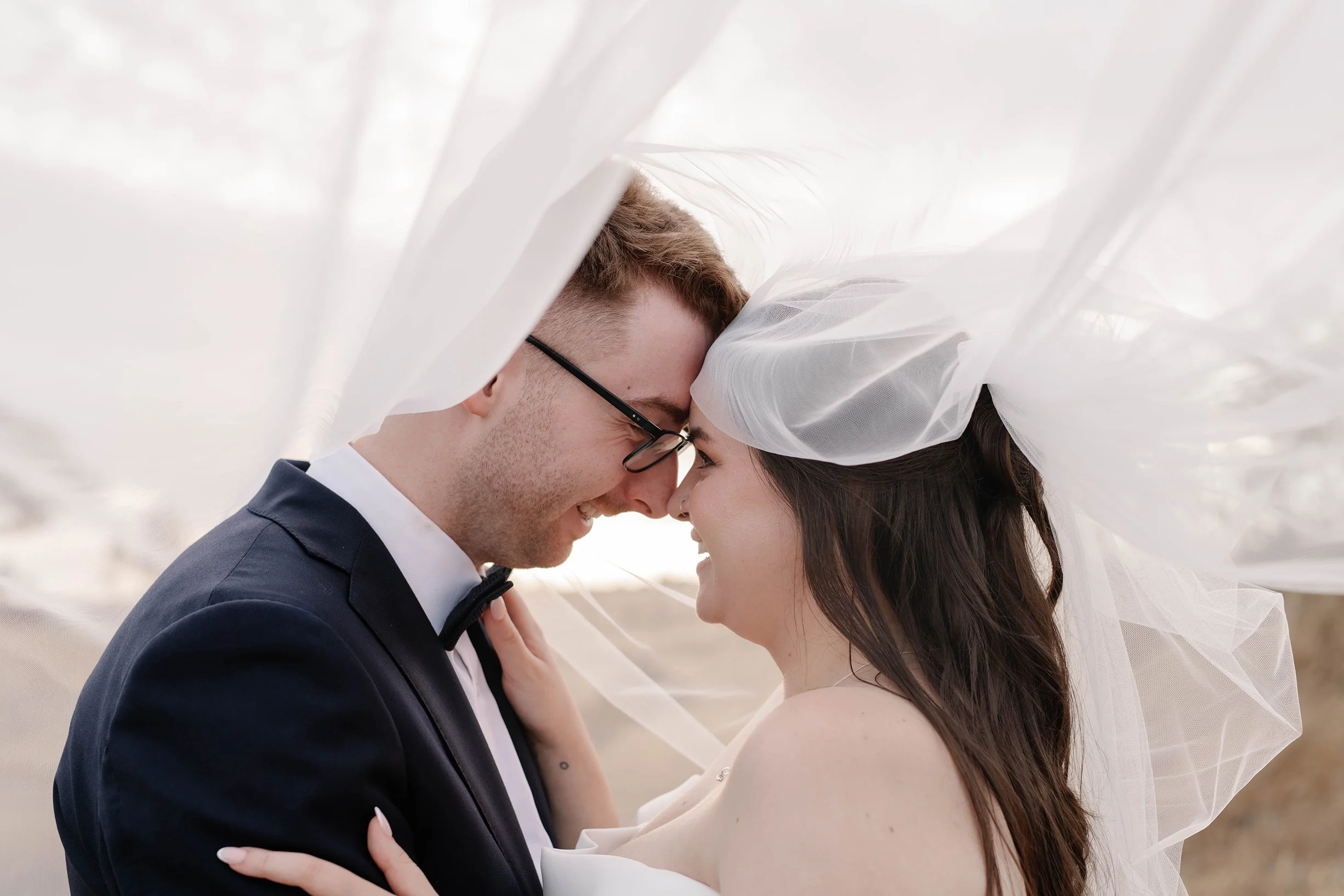 A bride and groom share a close, affectionate moment, their foreheads touching, under a sheer white veil during a wedding photoshoot.
