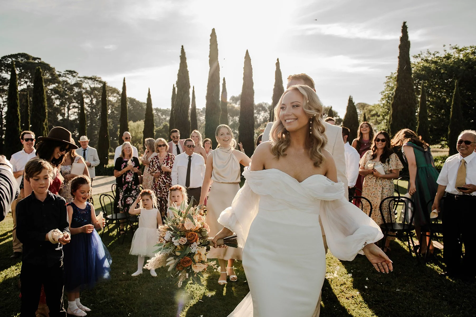 Bride beaming as she exits the outdoor ceremony aisle at Barn and Co wedding venue with cypress trees and golden sunlight in the background