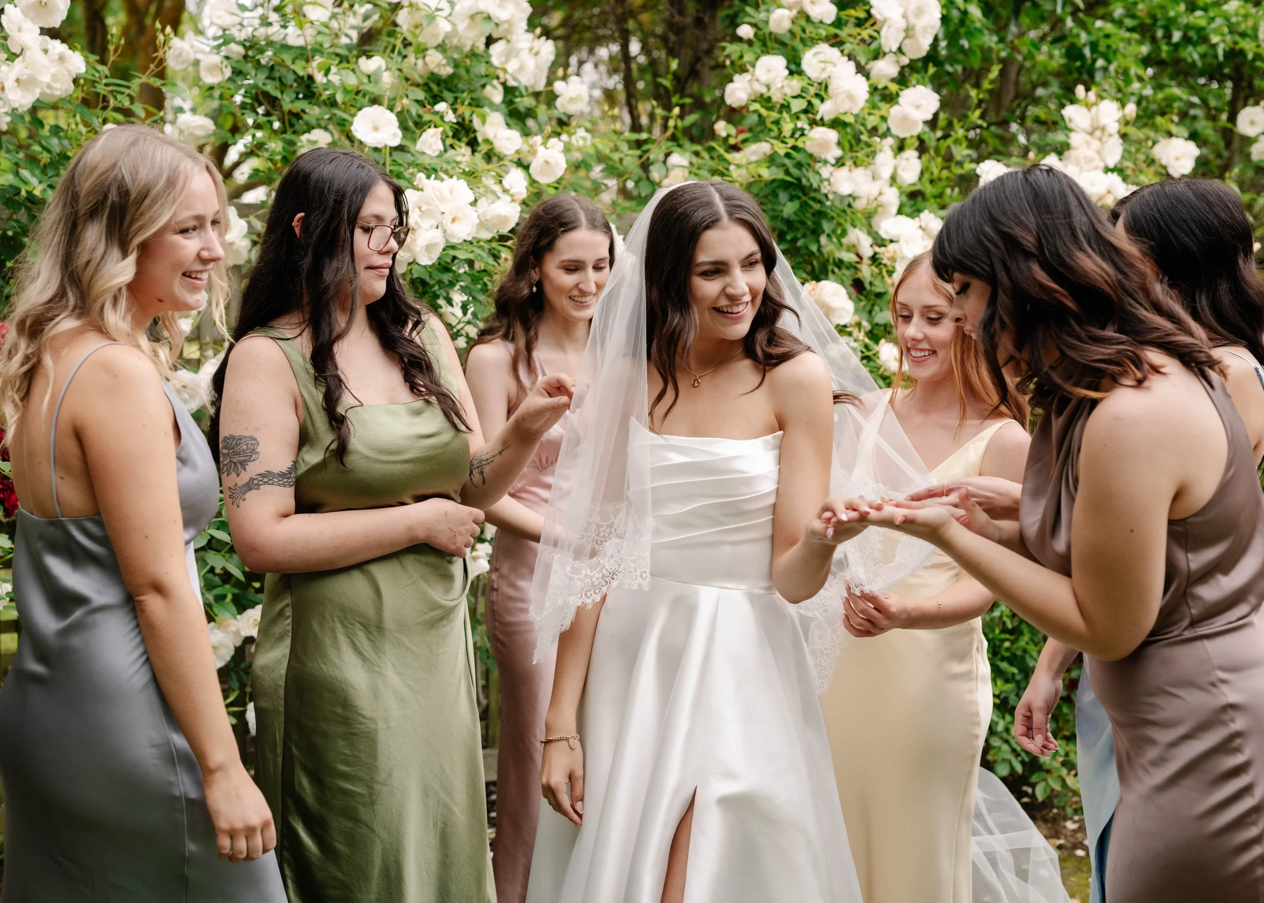 Bride and bridesmaids in mismatched satin dresses admiring the wedding ring in front of white rose bushes at Dalywaters Rose Garden Victoria