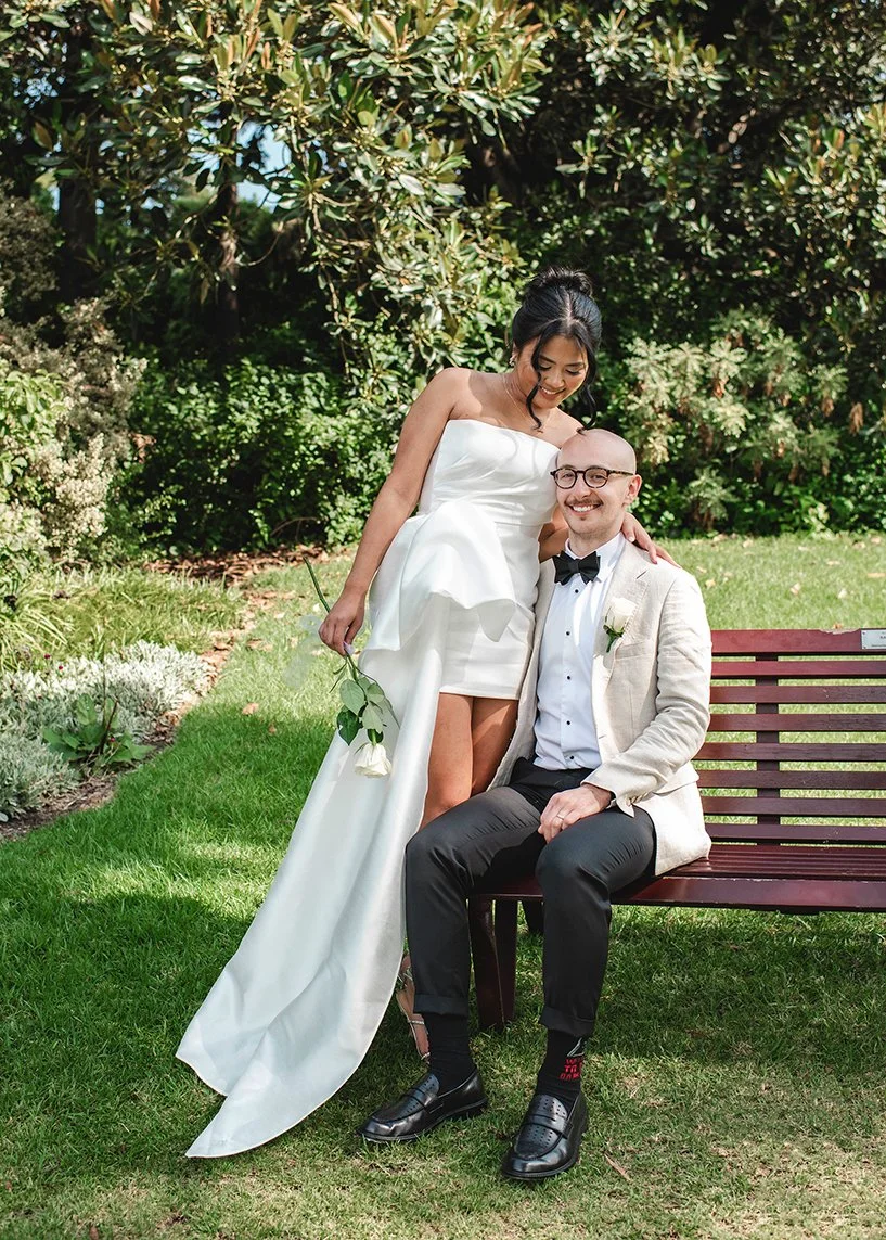 A bride in a white wedding dress and a groom in a beige blazer and black bow tie sitting on a park bench, surrounded by green trees and grass, smiling and holding a white flower.