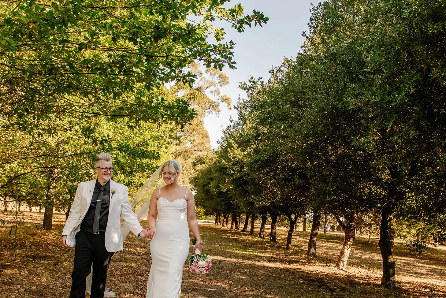 A newlywed couple walking in a green park holding hands, with trees and sunlight in the background.