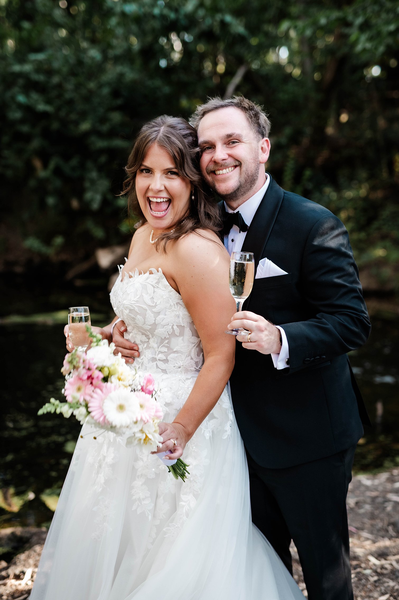 A smiling bride and groom in wedding attire celebrate outdoors, holding champagne flutes, with a bouquet of pink and white flowers, standing near a body of water surrounded by trees.