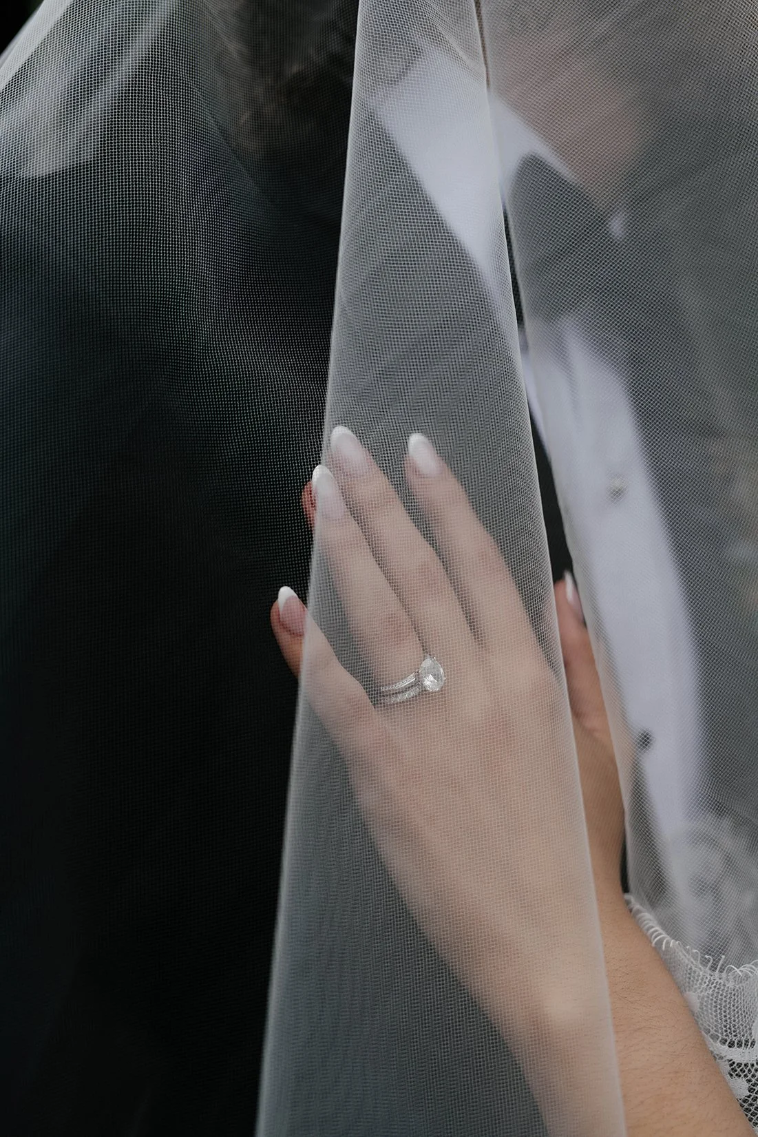 Close-up of a woman's hand with a large diamond engagement ring, gently touching a sheer white veil.