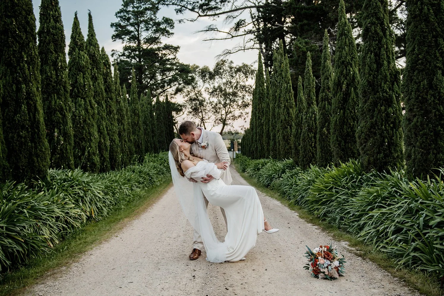 A bride and groom in wedding attire sharing a romantic kiss on a dirt path lined with tall, green cypress trees and lush foliage, with a bouquet of flowers on the ground nearby.