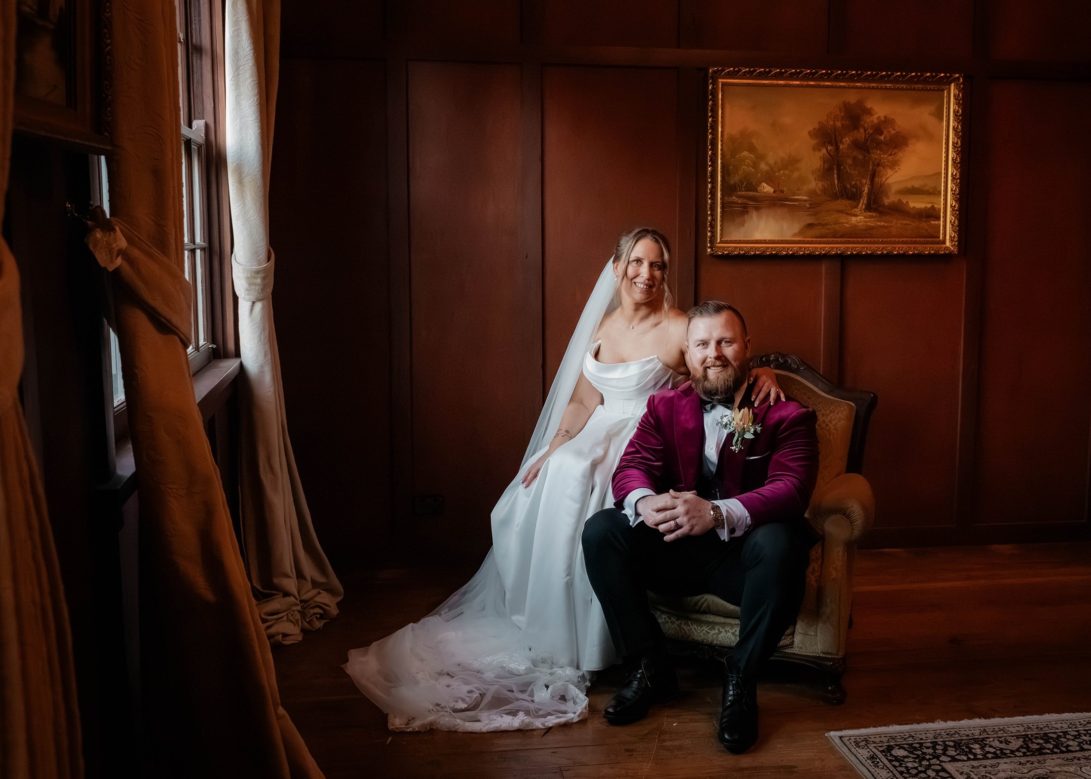 A bride and groom pose in a dimly lit room with wooden walls and a framed landscape painting. The bride is standing and wearing a white wedding dress with a veil, while the groom is seated, wearing a burgundy velvet jacket with a boutonniere, and smiling at the camera.