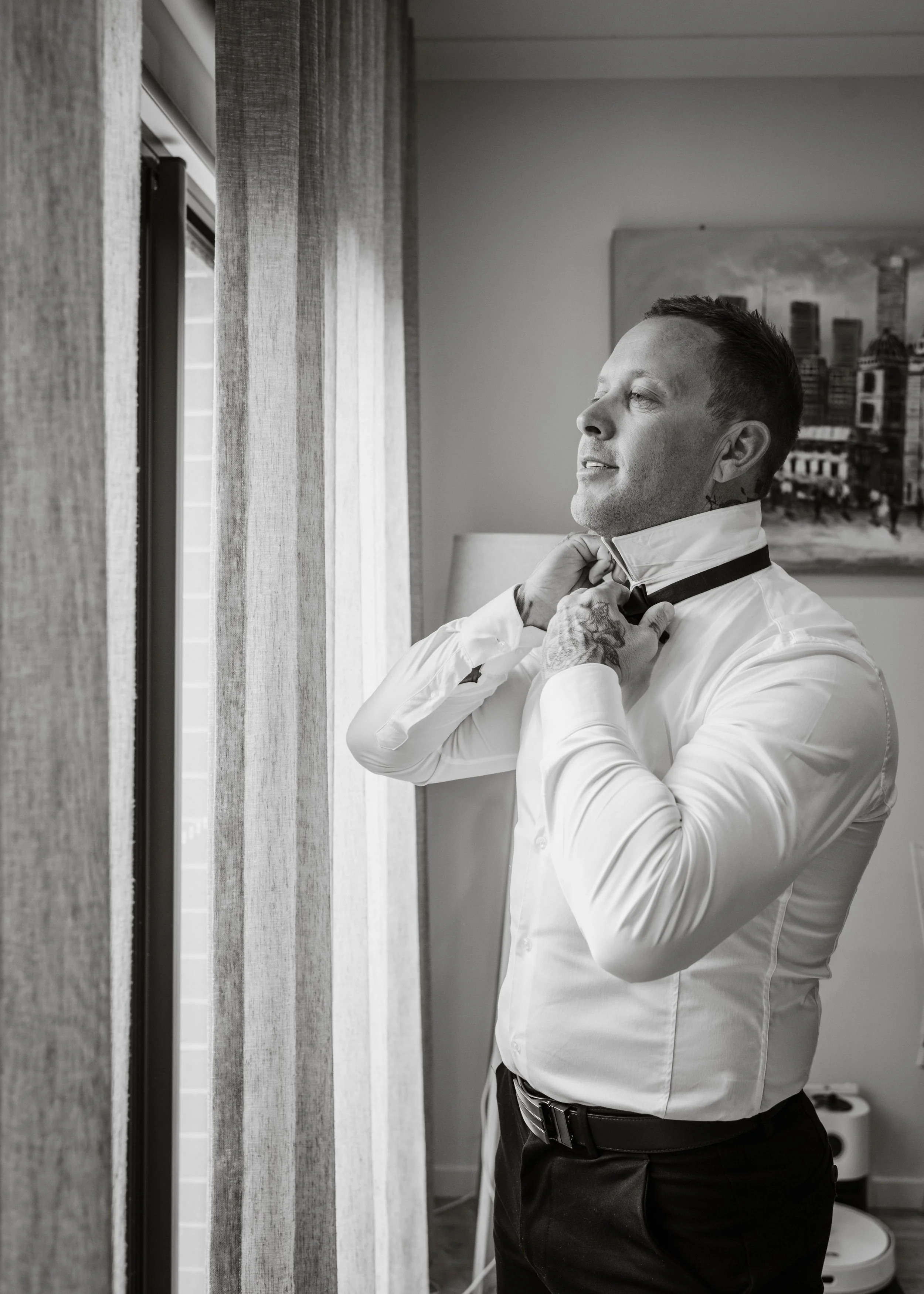 Groom adjusting his bow tie by a window while getting ready for his wedding at Jason's Restaurant Beaconsfield Victoria