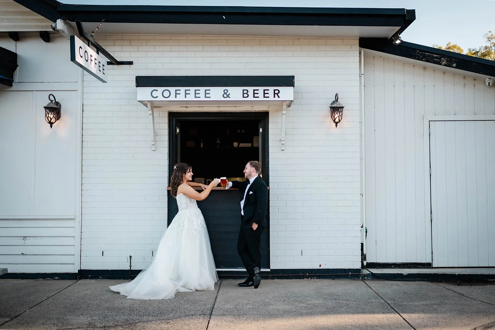 A newlywed couple in wedding attire outside a cafe that serves coffee and beer, toasting with glasses of beer.