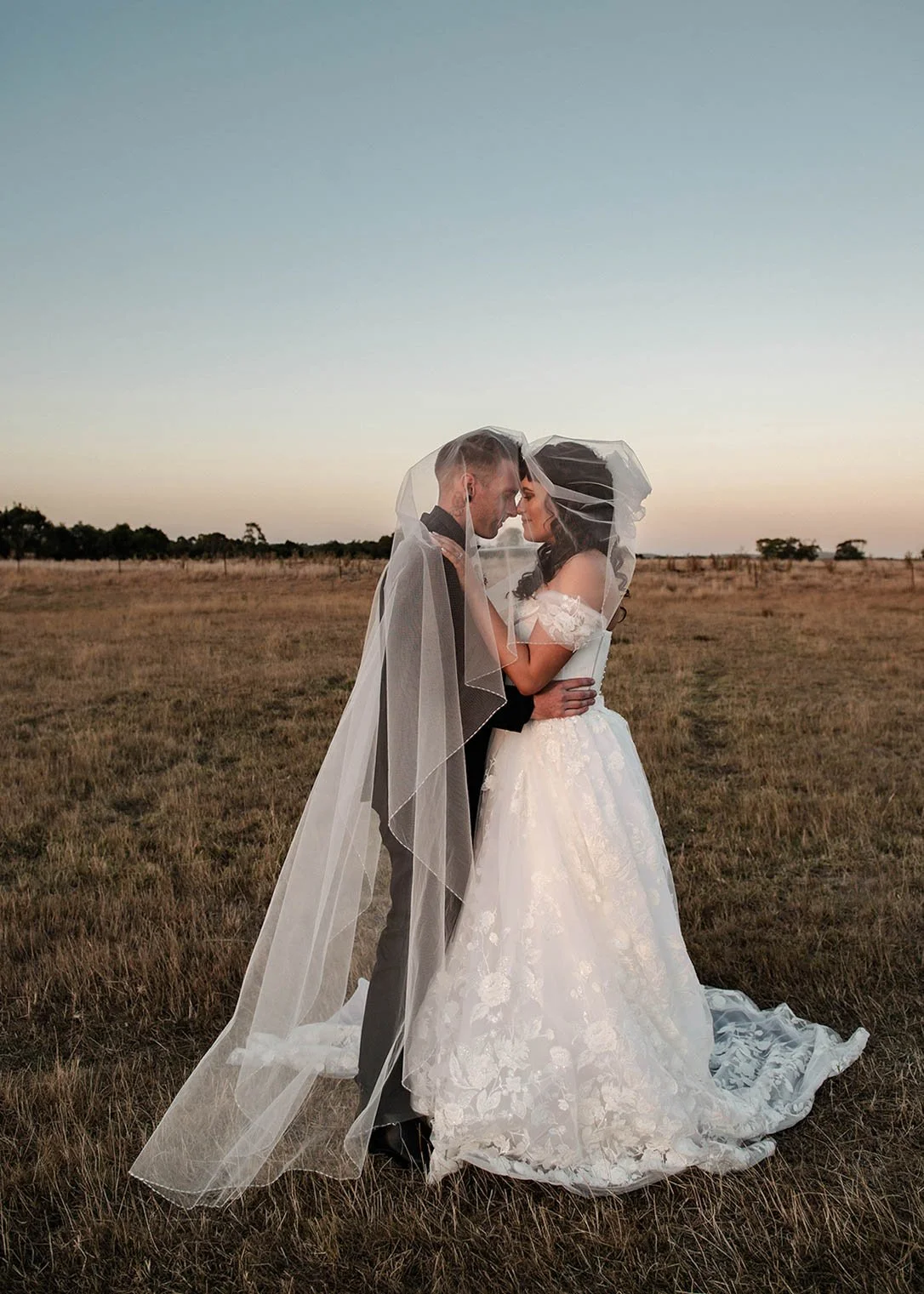 Bride and groom kissing beneath a long wedding veil in an open field at sunset