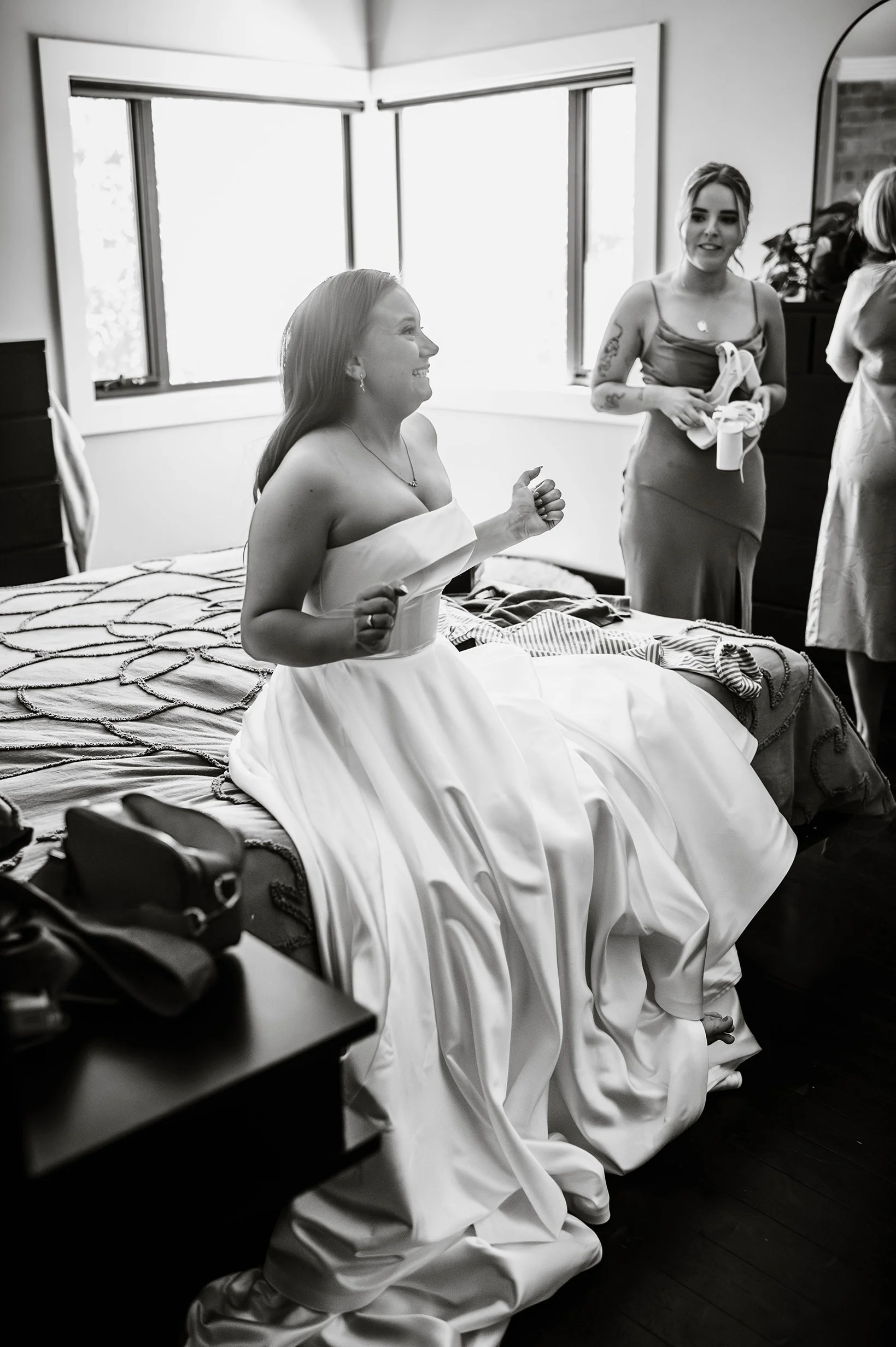 Candid black and white of bride in wedding dress sitting on bed excited