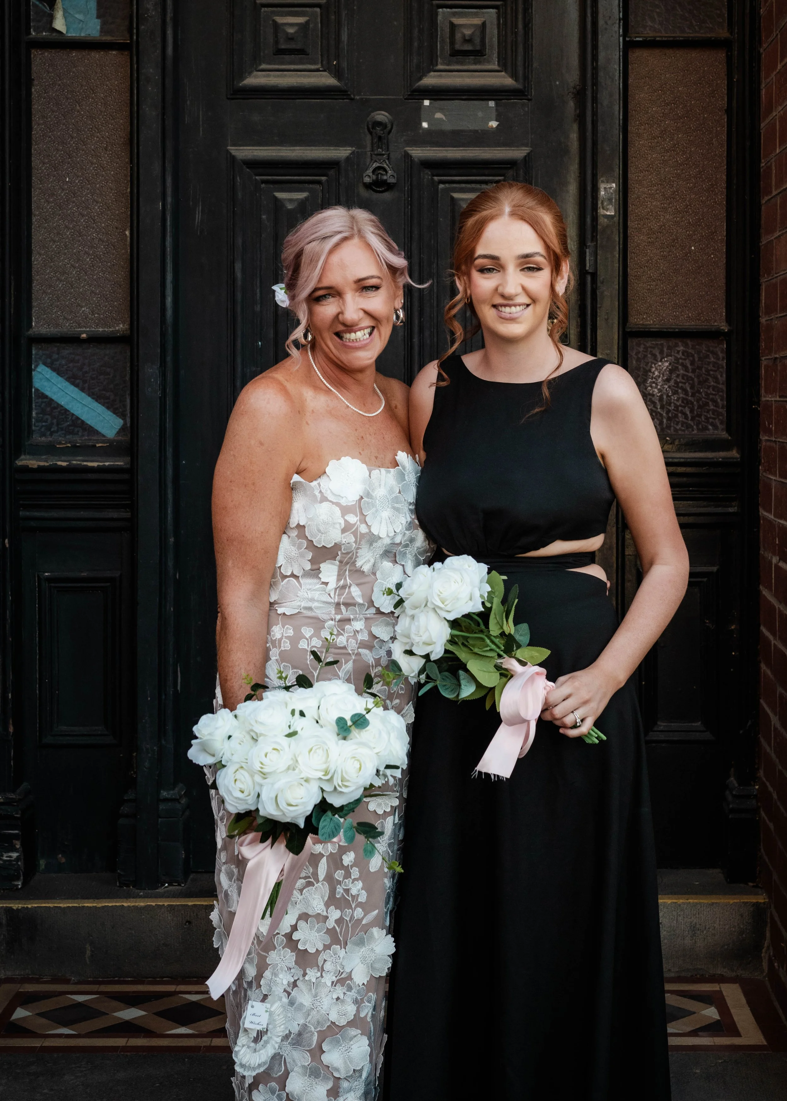 Bride in a floral wedding dress standing with a bridesmaid in a black dress holding white bouquets in front of a dark doorway