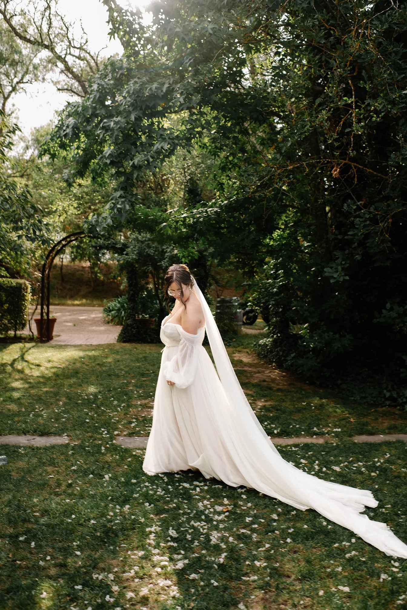 Bride looks down at dress trailing through white petals on lit Chateau Dore garden grass in Bendigo