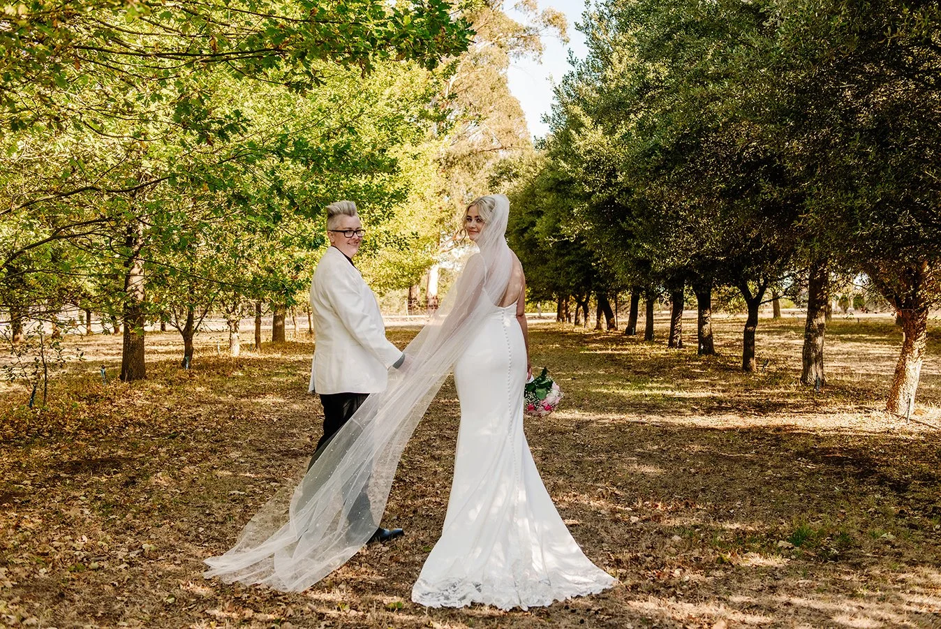 A bride in a white wedding gown and veil holding a bouquet and a person in a white suit holding her hand in a wooded outdoor setting.