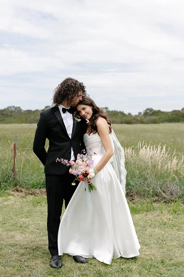 Bride and groom standing in a field, with the groom dressed in a black tuxedo and the bride in a white wedding dress holding a bouquet of flowers.