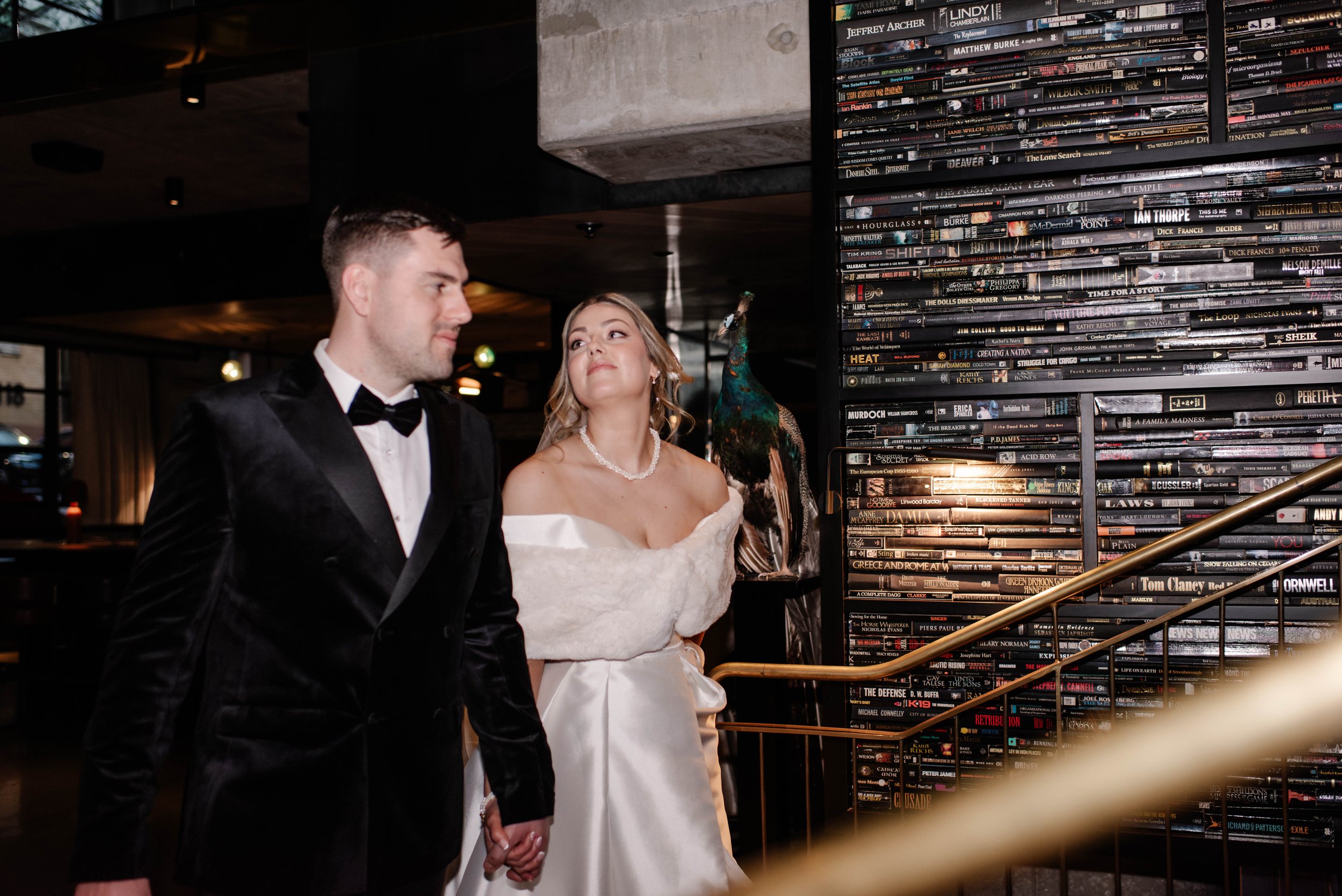 A bride and groom in wedding attire holding hands and walking inside a building with a wall of magazines or books in the background.