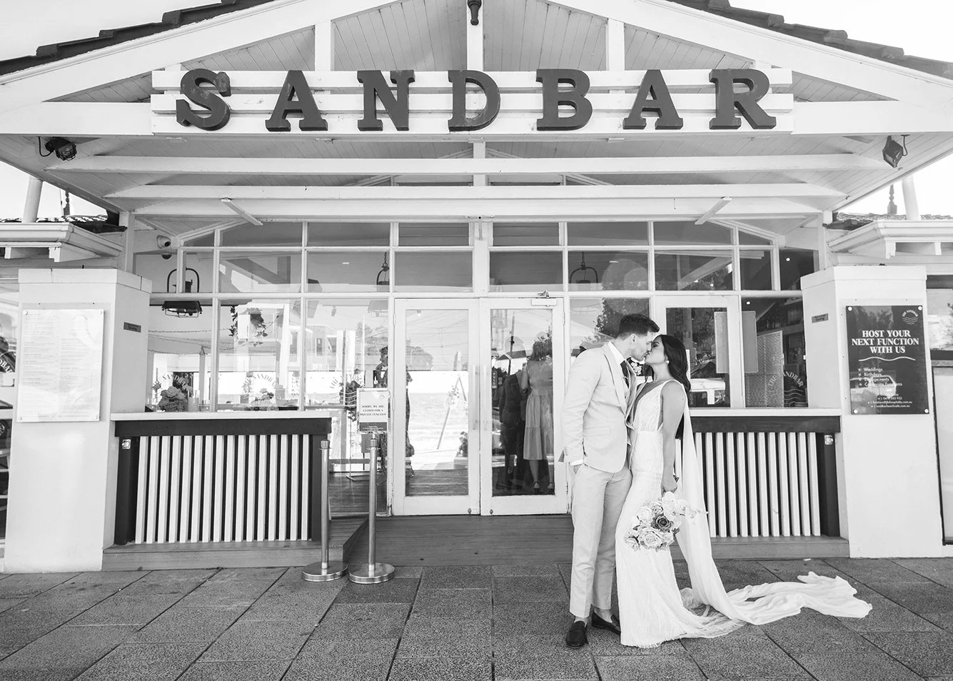 A couple in wedding attire kissing outside a sandbar restaurant, with the bride holding a bouquet of flowers, in front of glass doors and windows, in black and white.