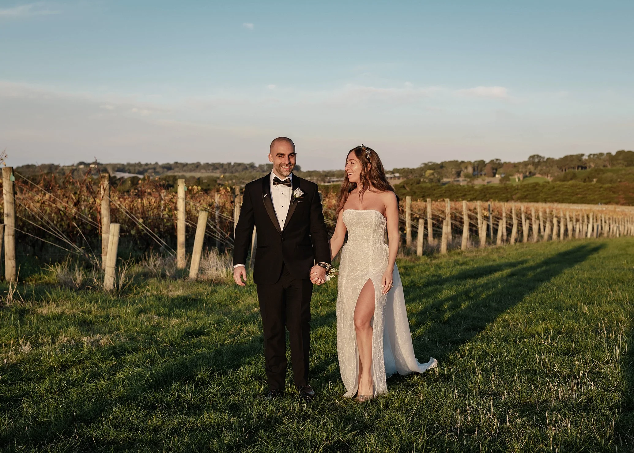 A newlywed couple walking hand in hand through a vineyard during golden hour, with a man in a black tuxedo and a woman in a strapless white wedding dress with a high slit.