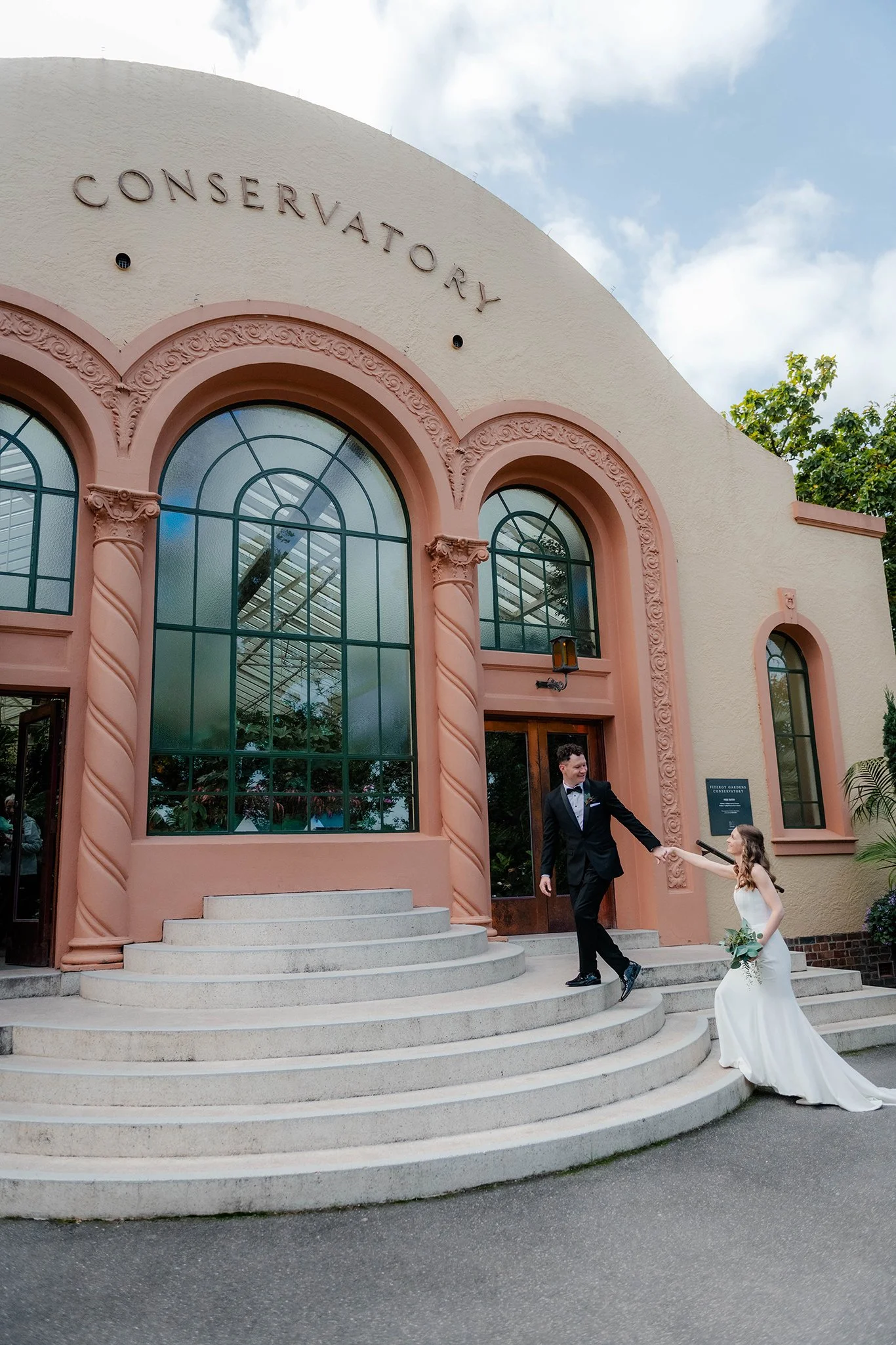 A man in a tuxedo and a woman in a white dress holding hands and walking down the steps of a building with a vintage look and arched windows labeled 'Conservatory'.