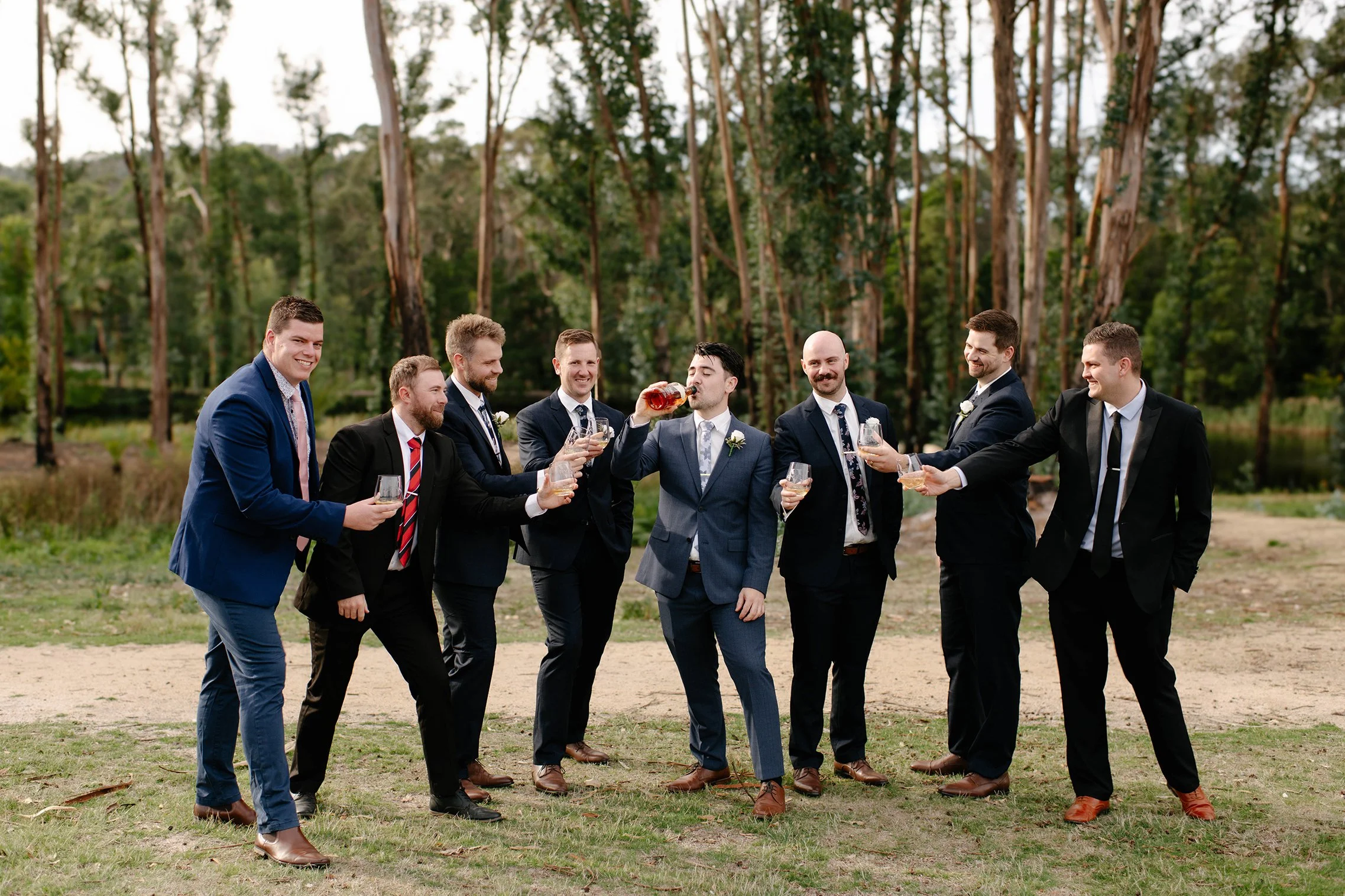 A group of men in suits standing outdoors, celebrating with drinks in a natural setting with trees in the background.