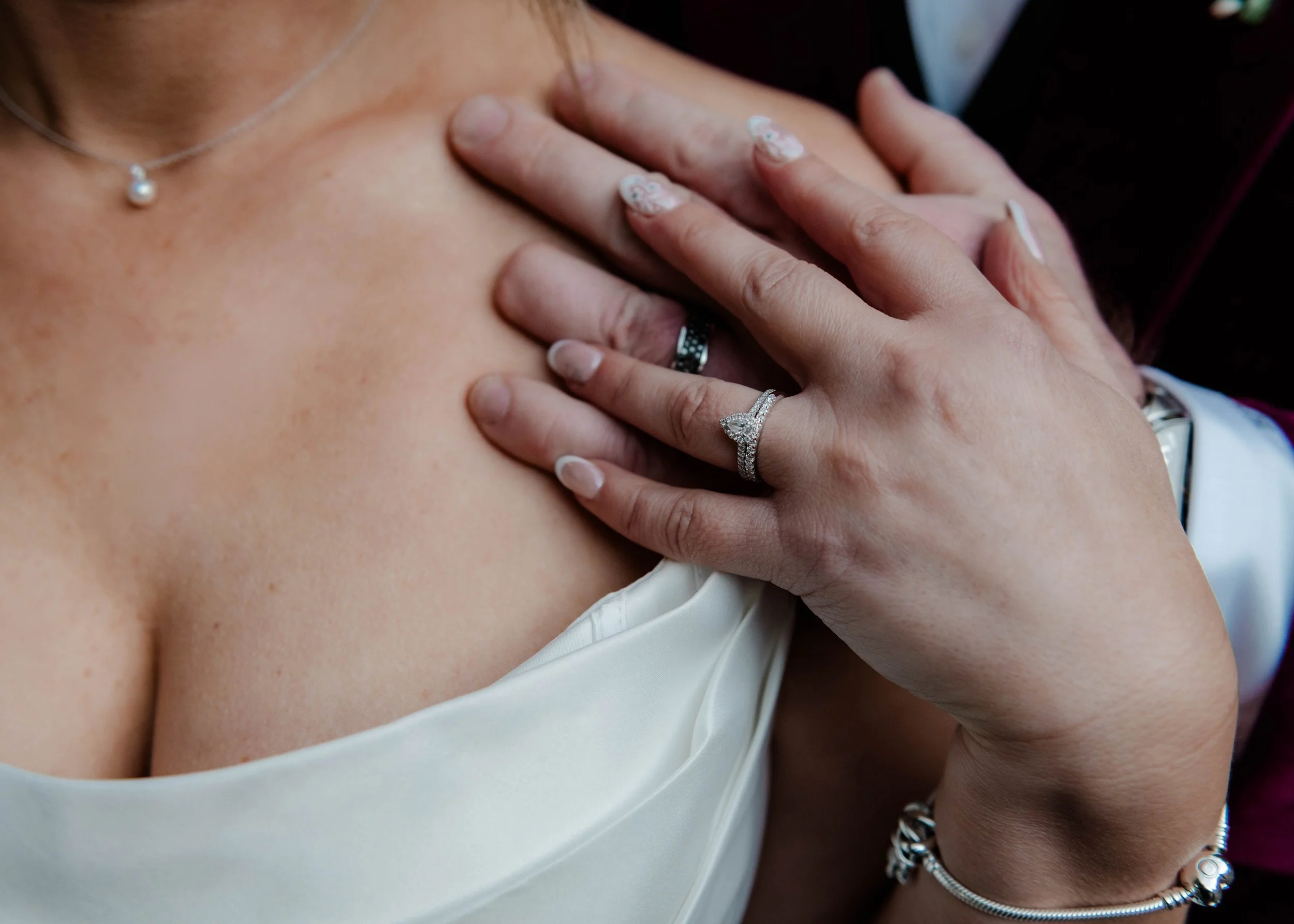 A woman wearing a pearl necklace, multiple rings including a diamond engagement ring, and a bracelet, gently touching her chest with her hand, dressed in a white top.