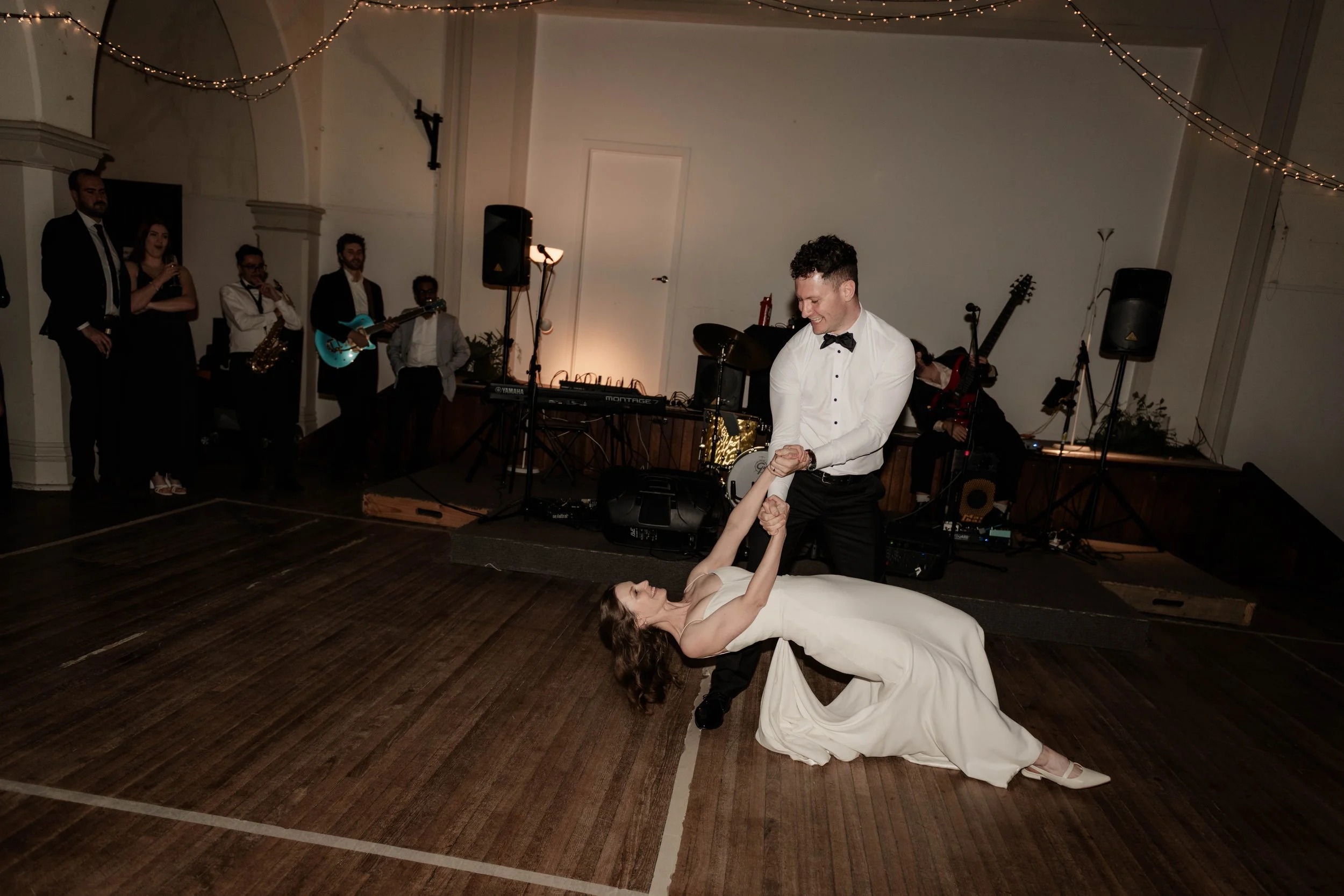 A wedding reception with a band playing, a groom and bride dancing in the center, the groom holding the bride's hand while she is lying on the dance floor in a white gown, and guests watching in the background.