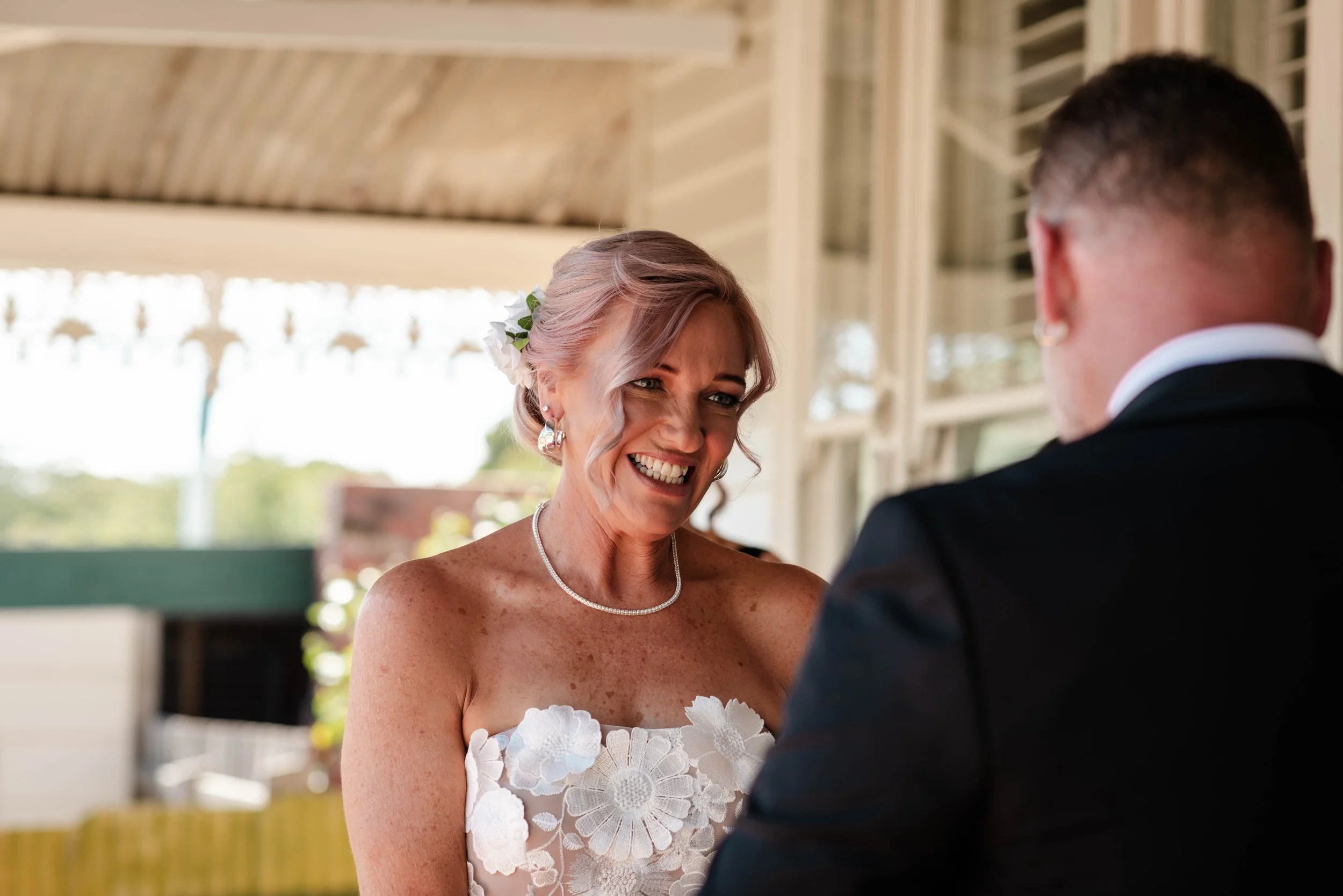 Bride in floral strapless wedding dress smiling at groom during an outdoor wedding ceremony on a covered veranda