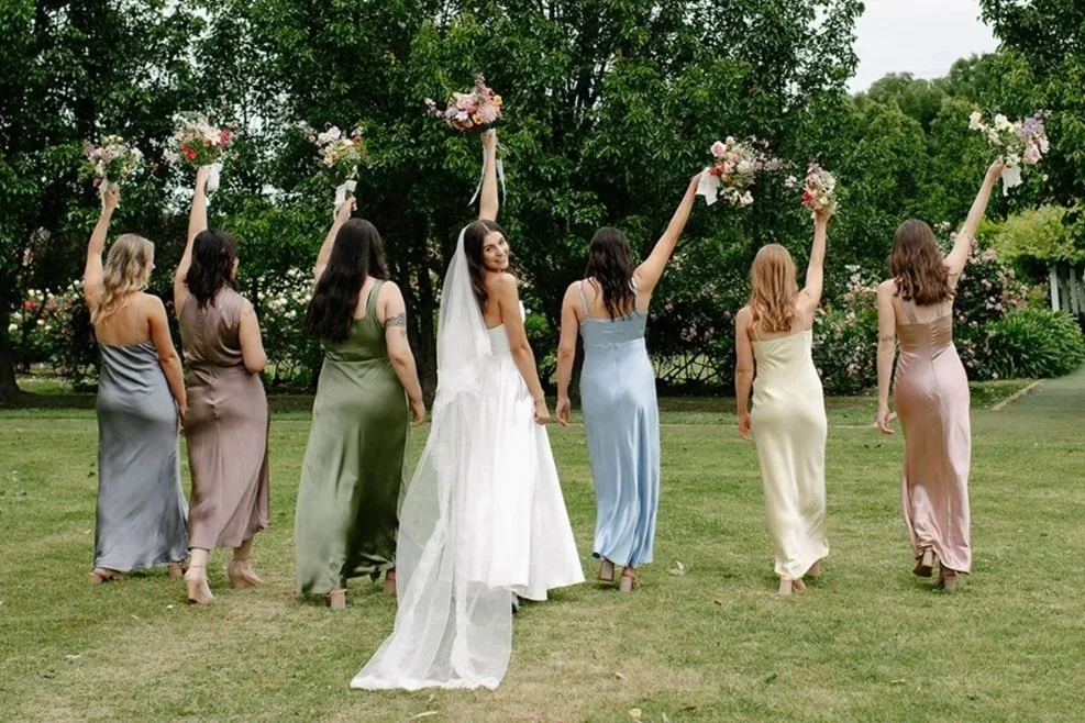 A bride in a white wedding dress standing outdoors with seven bridesmaids in pastel dresses, all holding bouquets and walking away from the camera in a lush green garden.