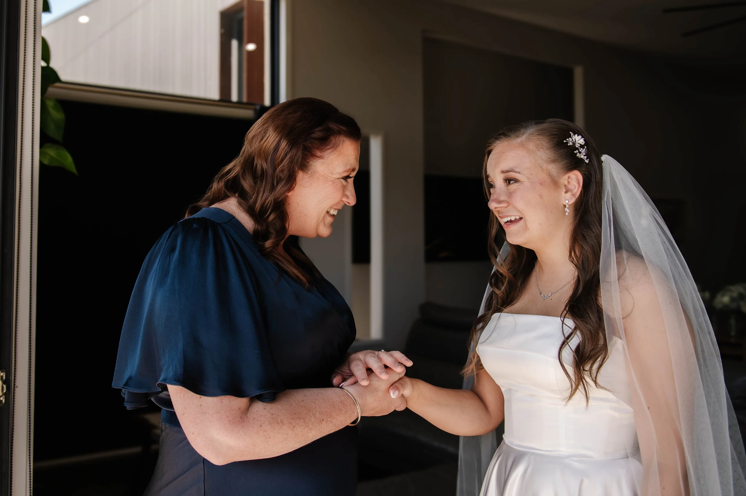 Mother of the bride clasps daughters hand as they both smile full of emotion