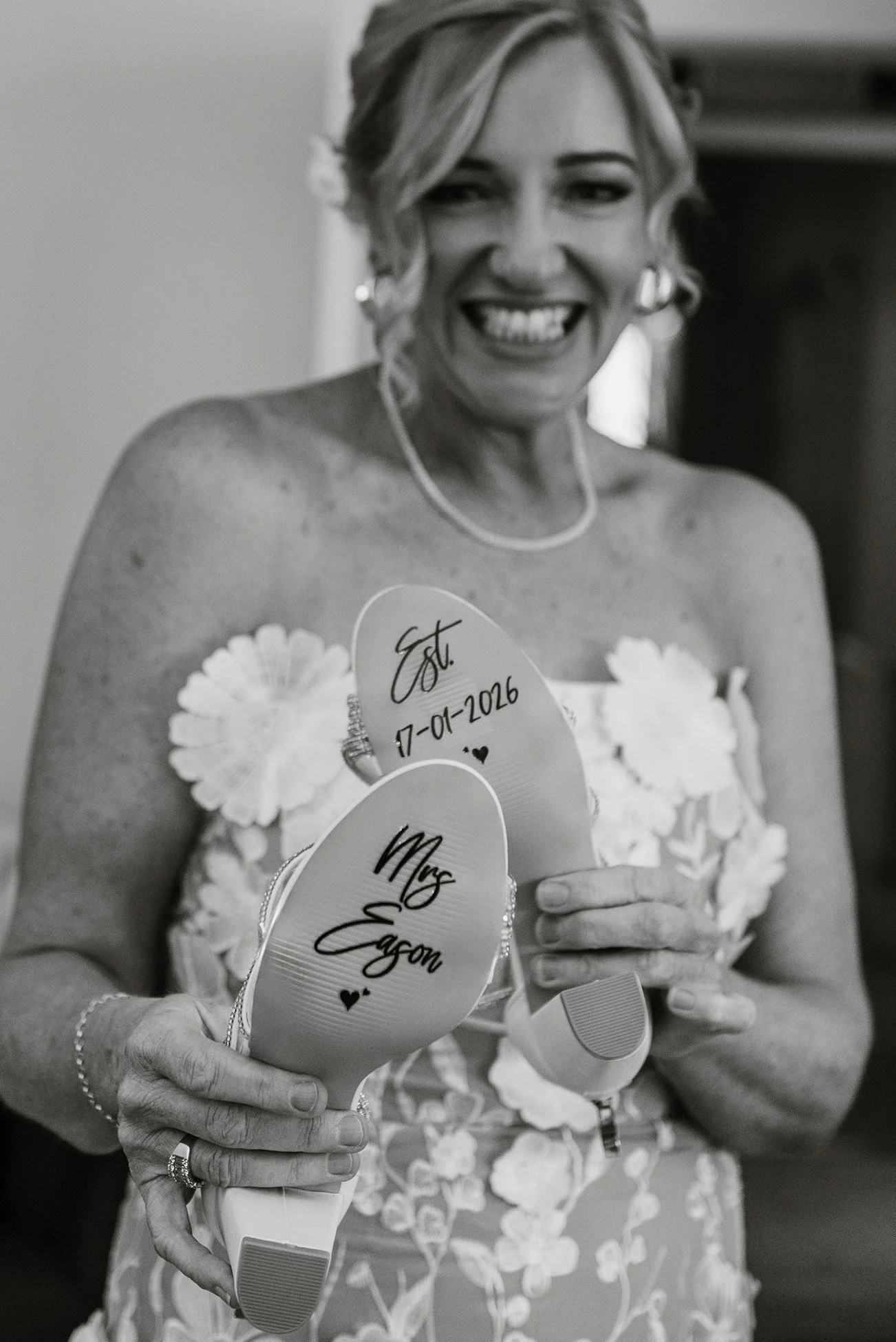 Black-and-white portrait of a smiling bride in a floral wedding dress holding up shoes with handwritten messages on the soles.