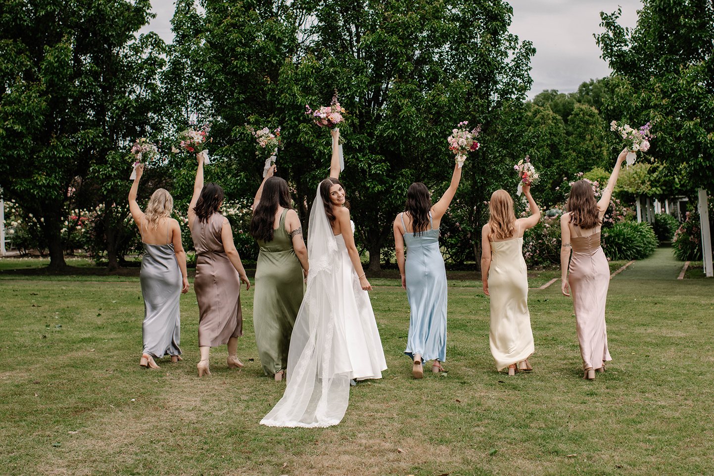 Bridal party outdoors with seven women, the bride in a white dress with veil in the center, and six bridesmaids in pastel dresses, holding bouquets of pink and white flowers, on a grassy area with trees in the background.