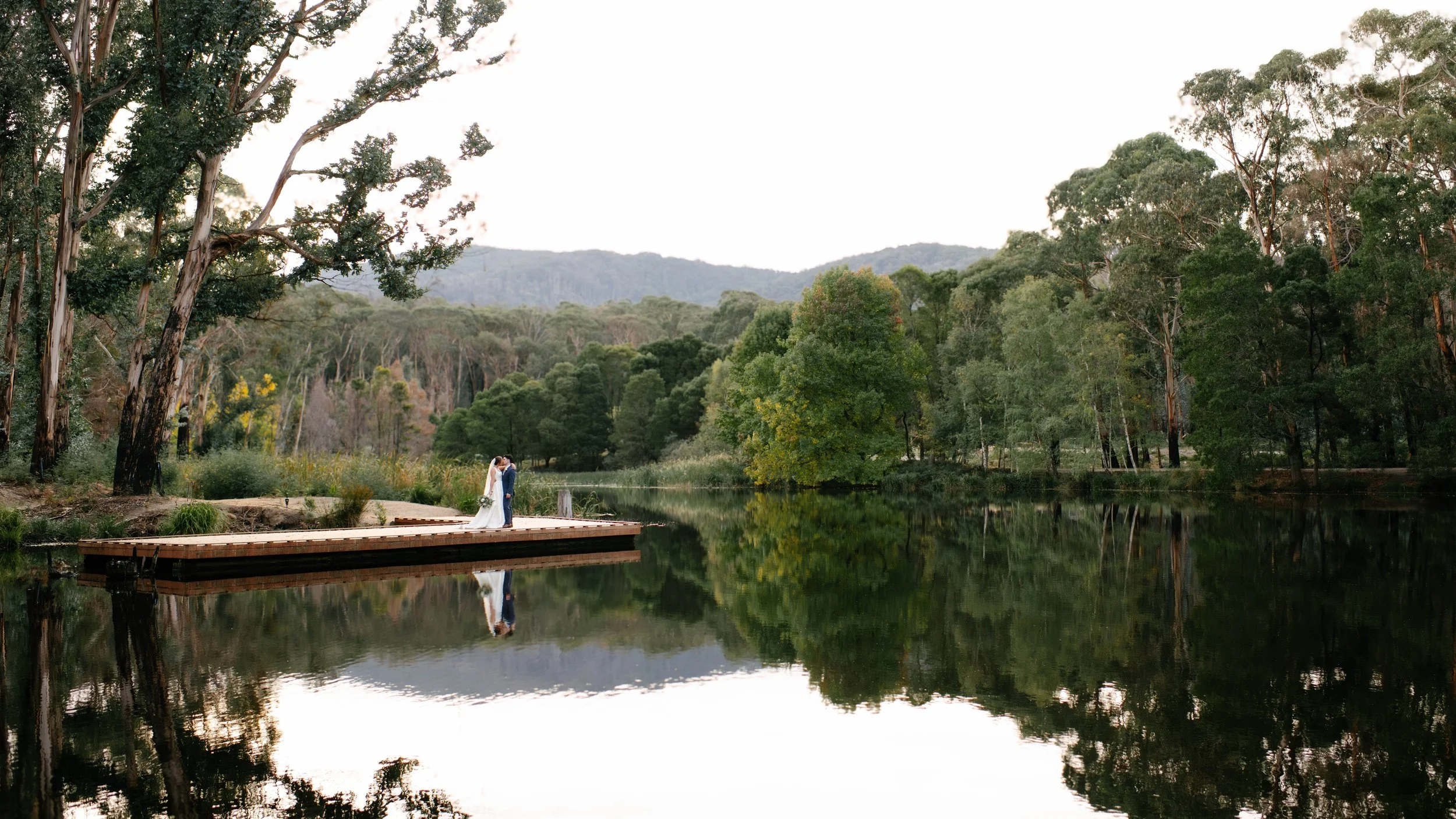 Bride and groom standing on a dock beside a calm lake with trees and mountain reflections in the background