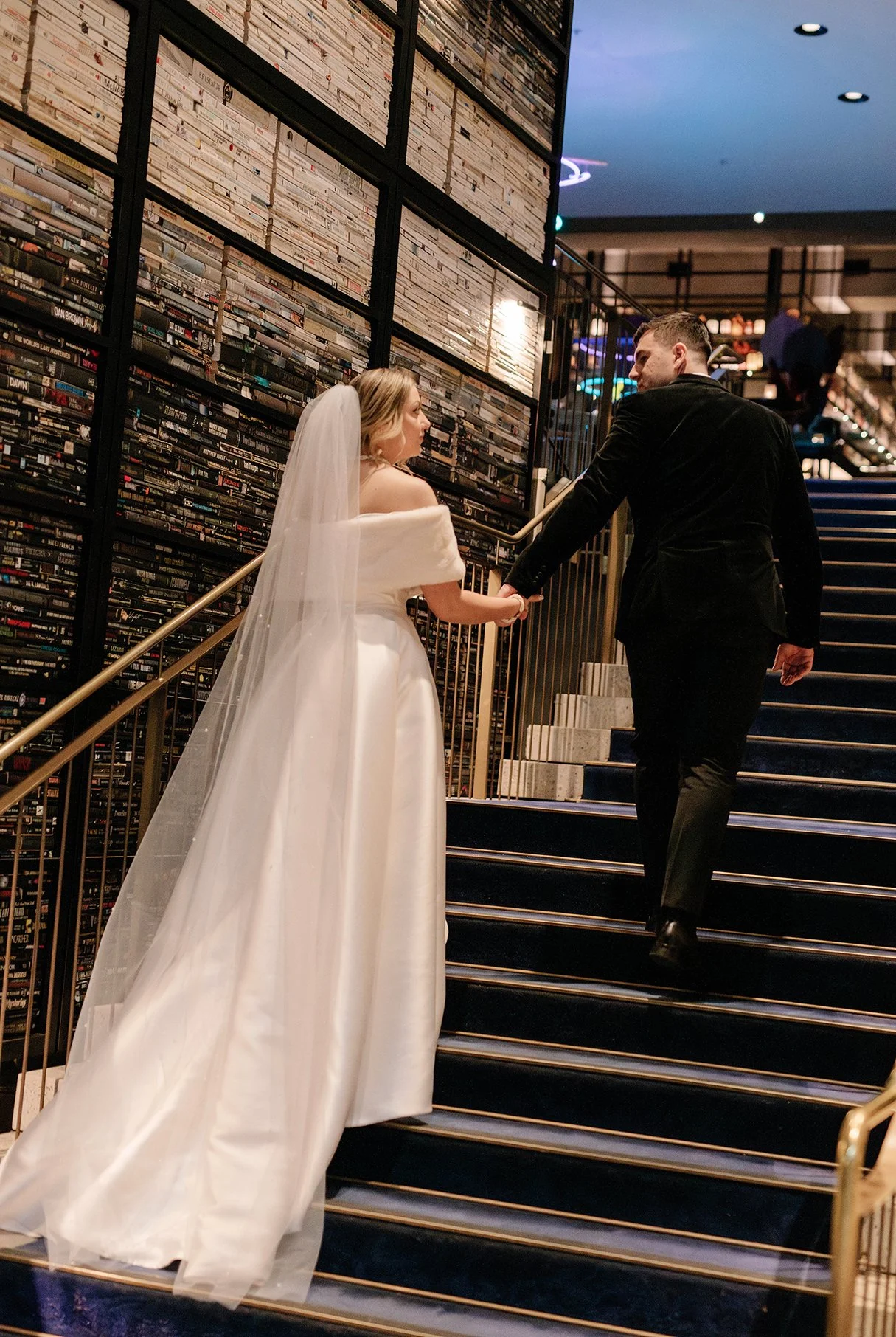 A bride in a white wedding dress holding hands with a groom in a black tuxedo walking up a staircase inside a venue decorated with a large collection of DVDs or books.