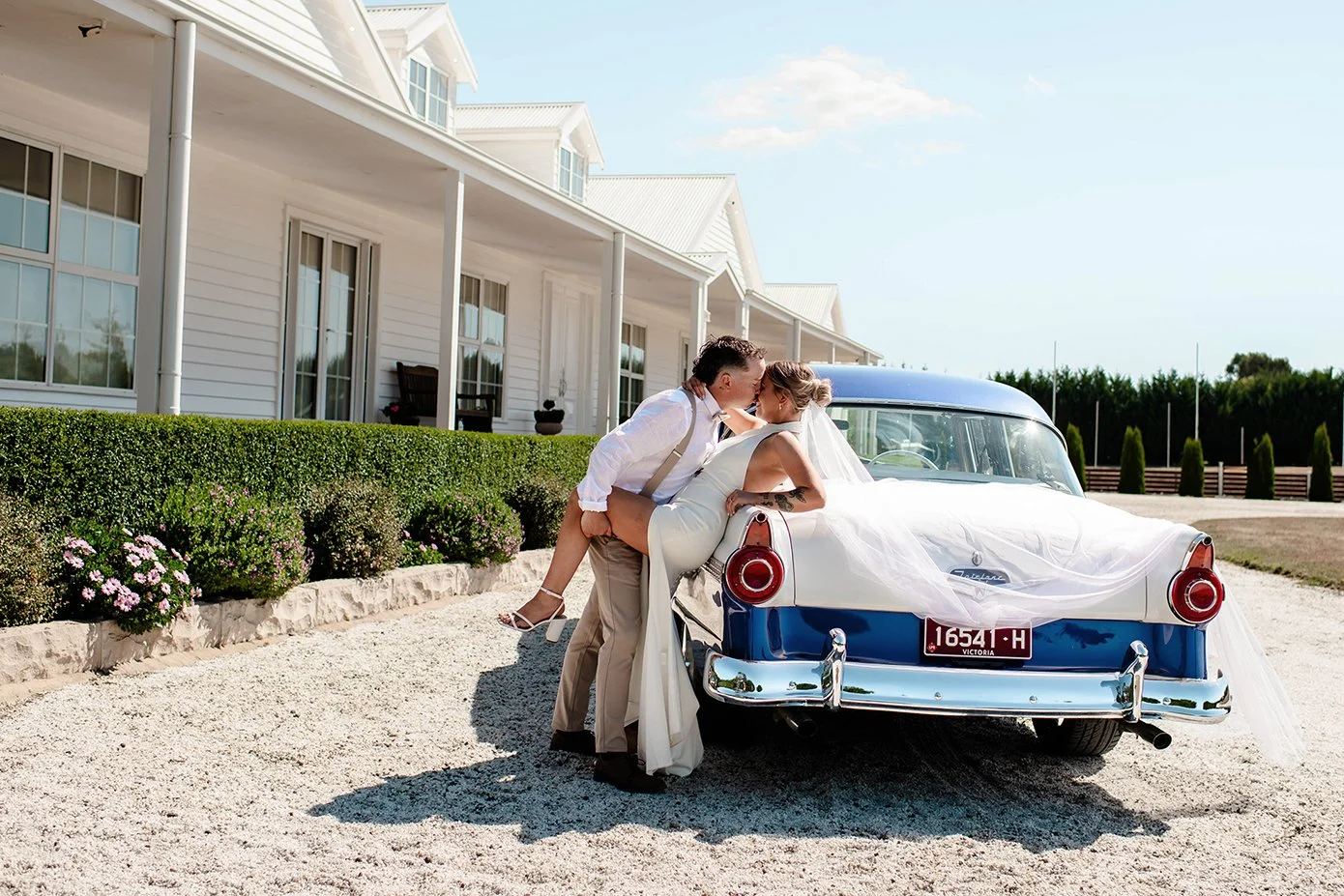 Bride and groom kissing on the back of a vintage blue American car outside a white country homestead at a Daylesford wedding