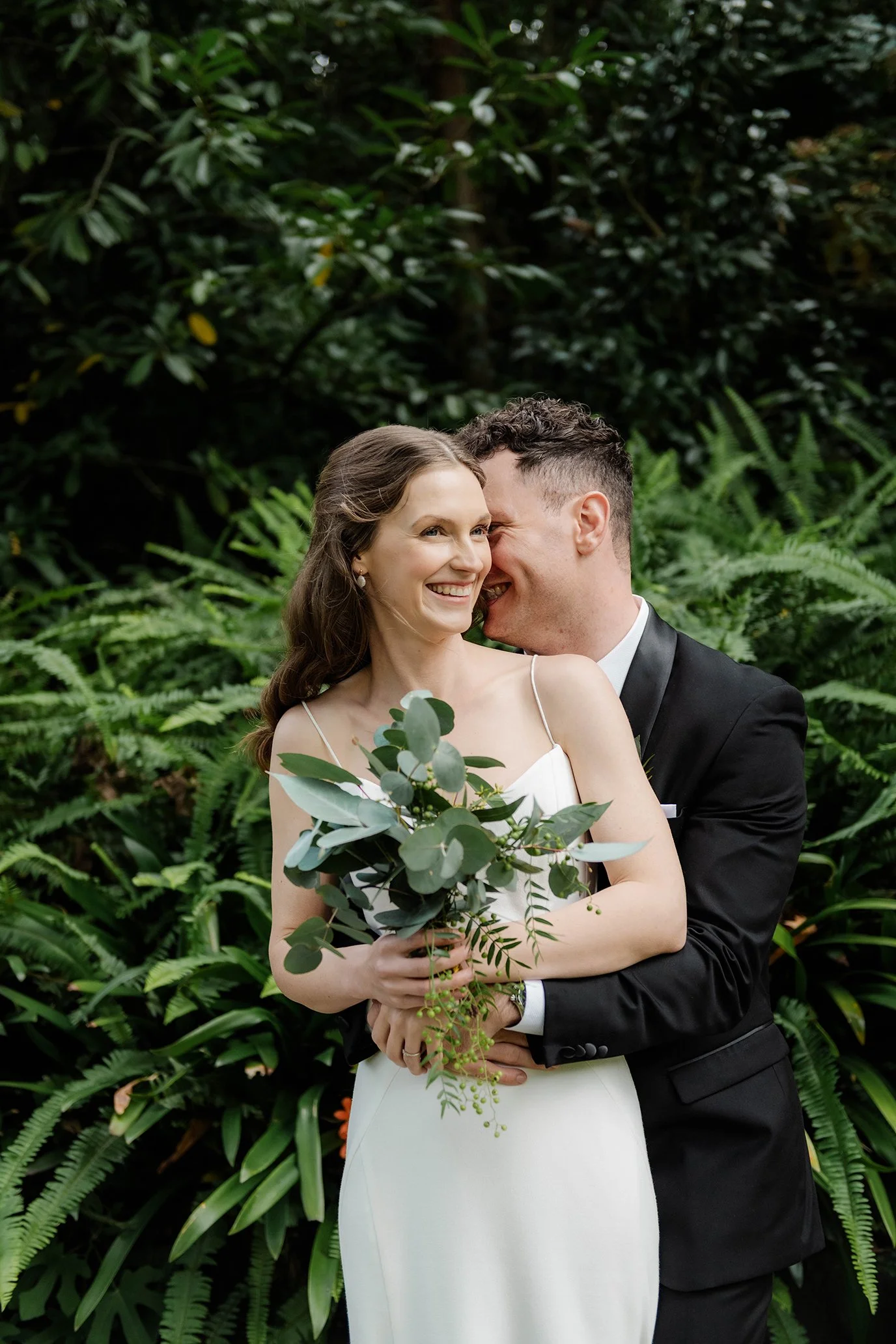 A bride and groom pose together outdoors, with the groom whispering into the bride's ear, surrounded by green foliage.