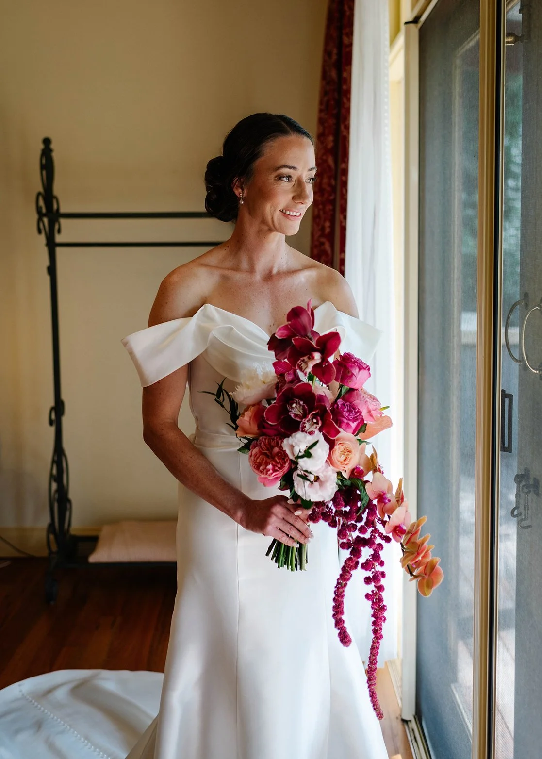 Bride standing by a window, holding a large bouquet of pink, red, and white flowers, dressed in an off-shoulder white wedding gown.