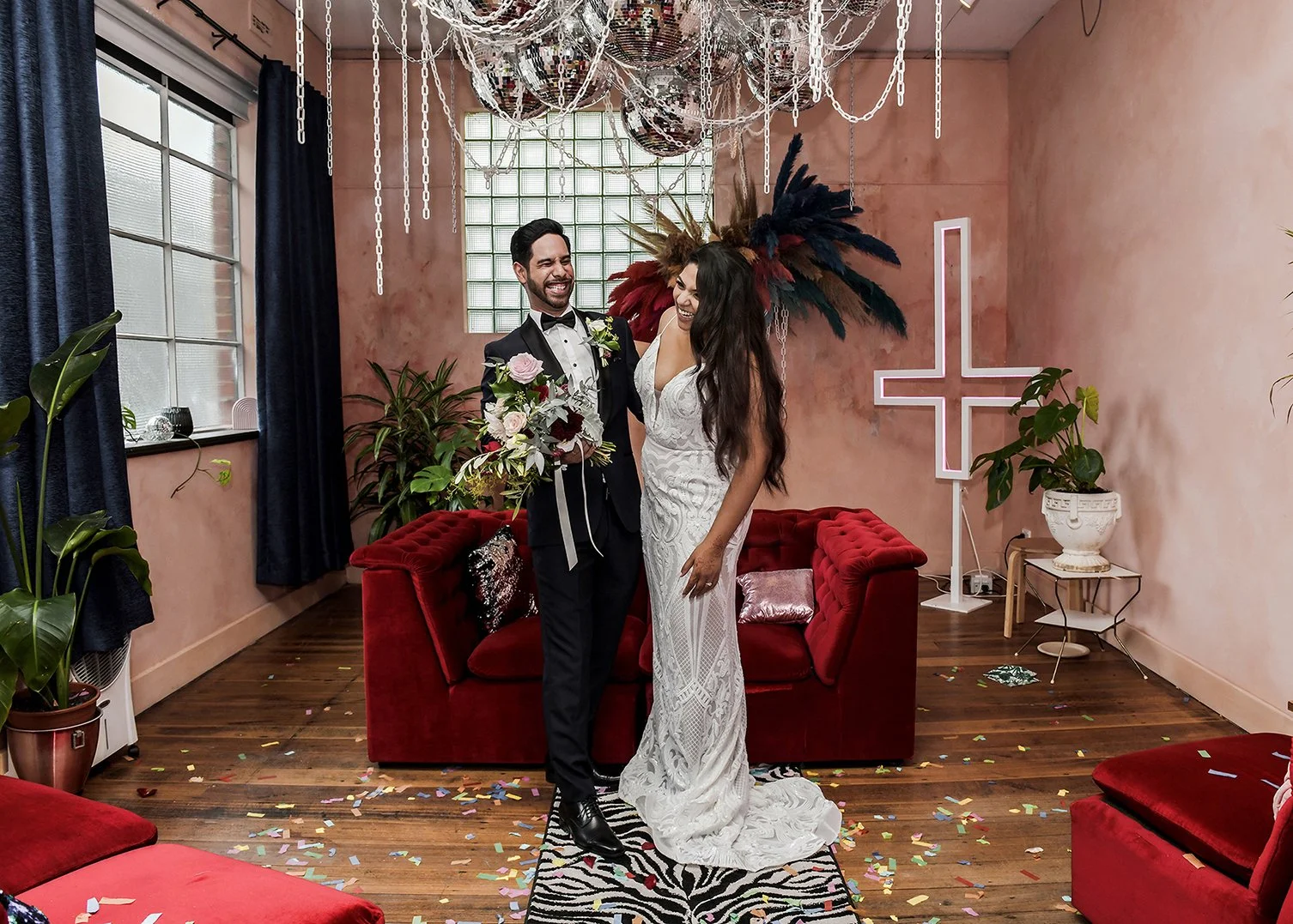 A bride and groom in wedding attire smiling and laughing together in a decorated indoor setting, with confetti on the floor, plants, and a neon cross on the wall behind them.