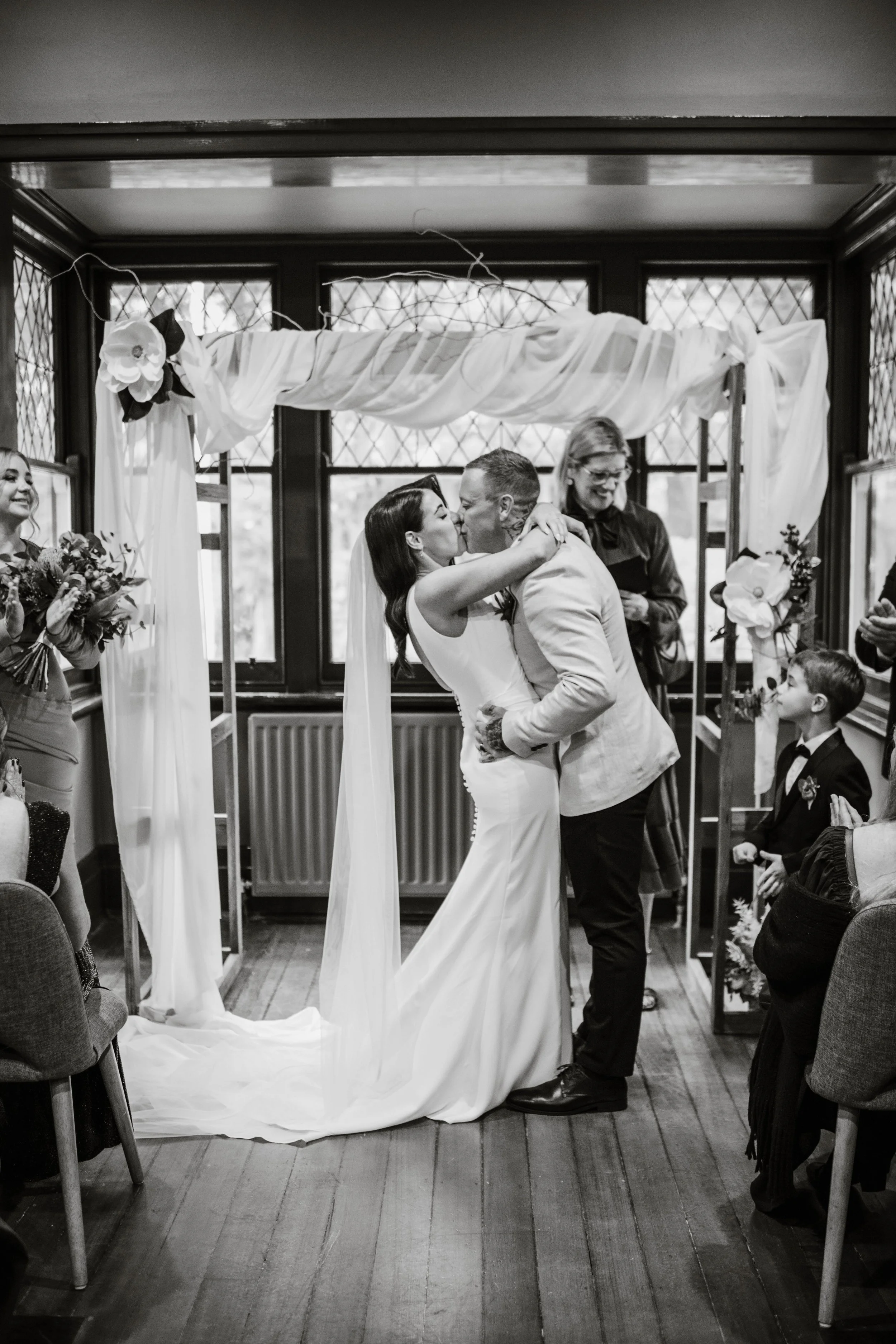 Bride and groom sharing their first kiss under a draped floral arch inside Jason's Restaurant Beaconsfield with guests cheering