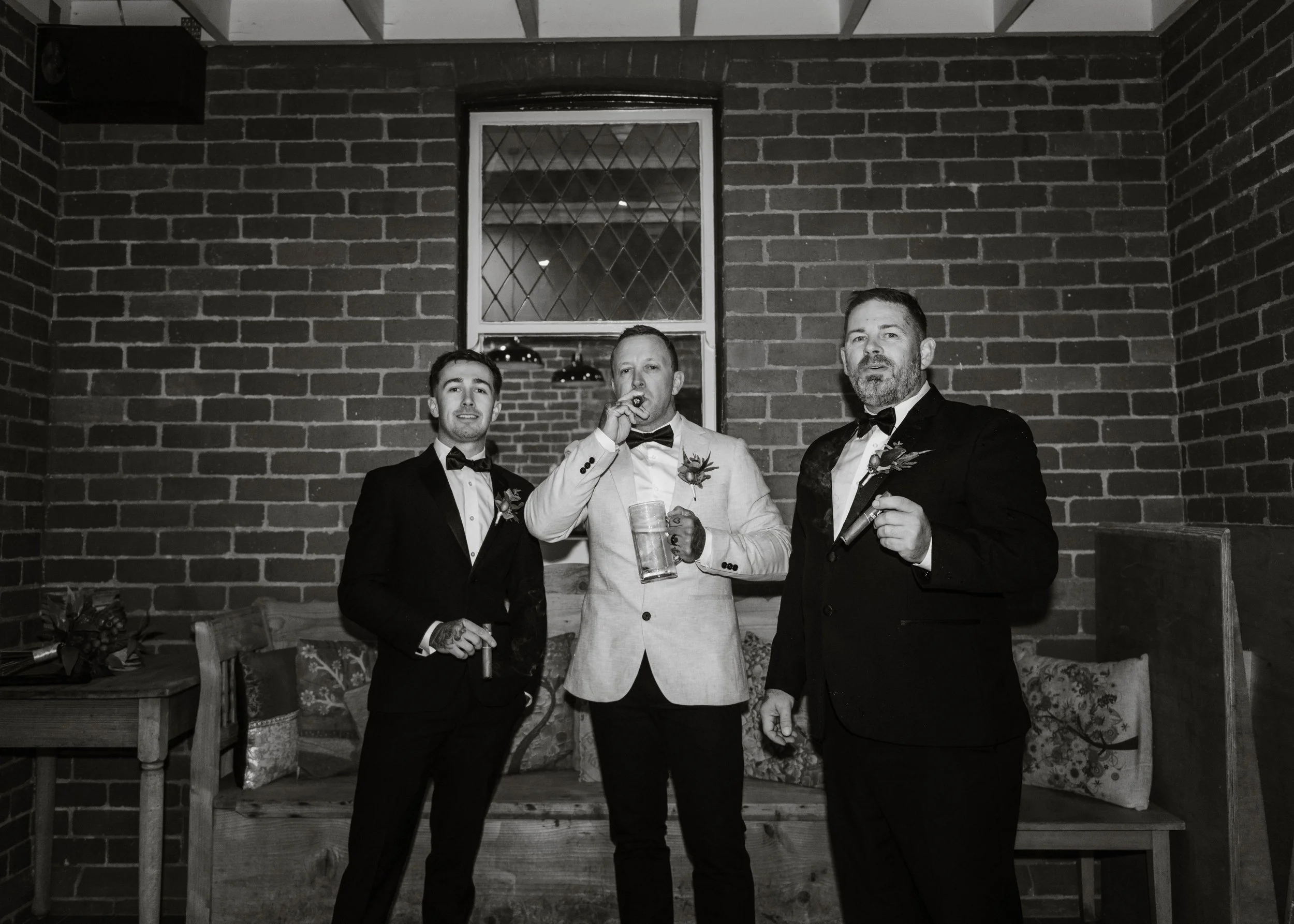 Groom and groomsmen celebrating with cigars and drinks against a brick wall at Jason's Restaurant Beaconsfield wedding reception