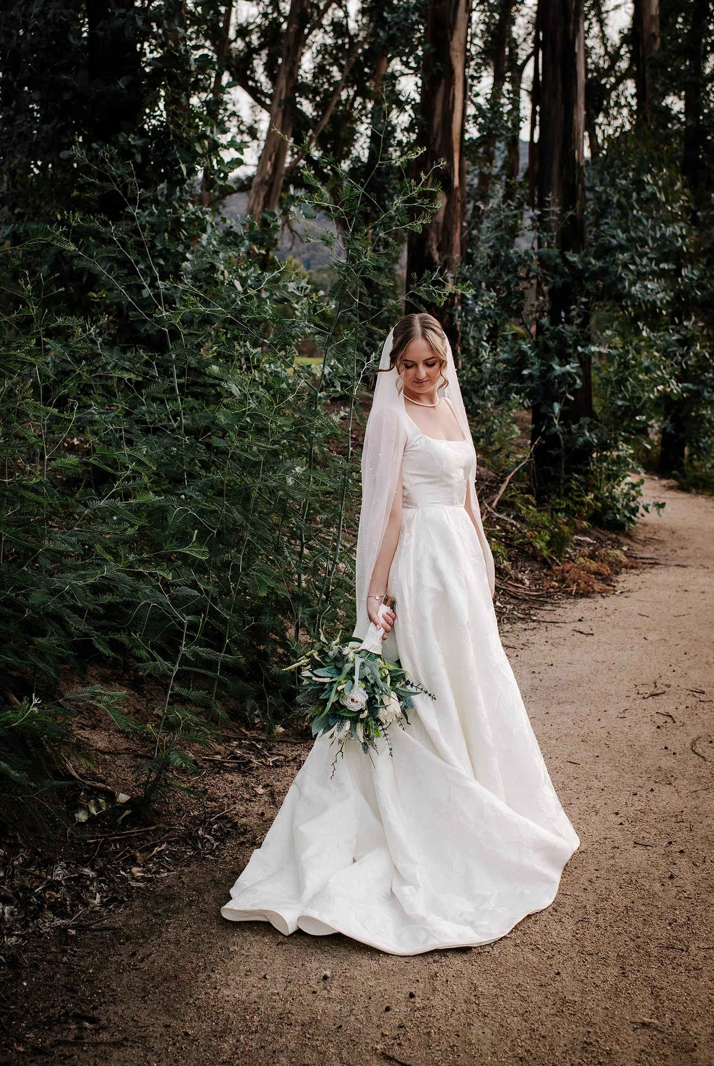 Bride in a white wedding dress holding a bouquet, standing on a dirt path next to green foliage and tall trees.