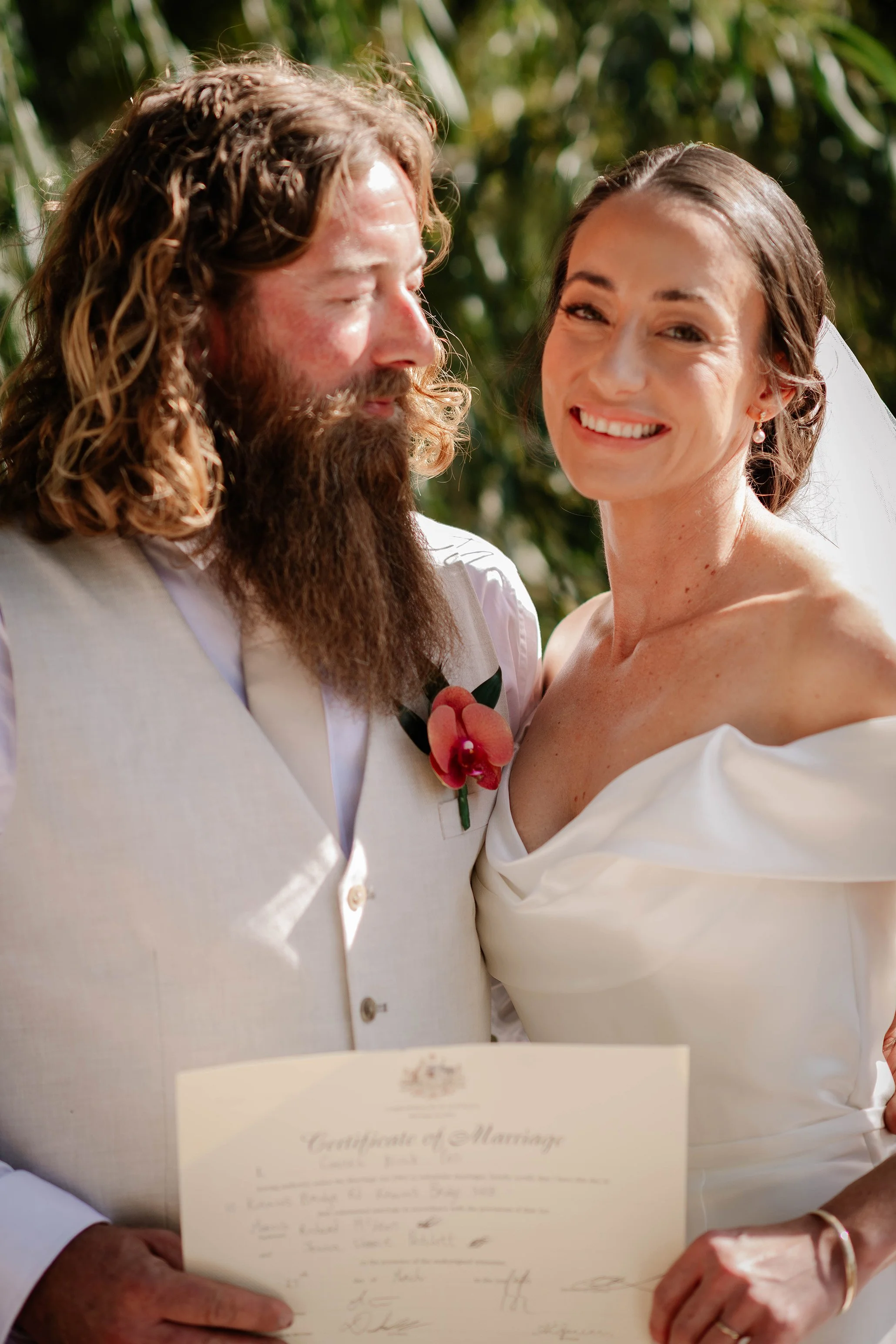 A smiling bride and a groom with long hair and a beard, holding a marriage certificate, standing outdoors with greenery in the background.