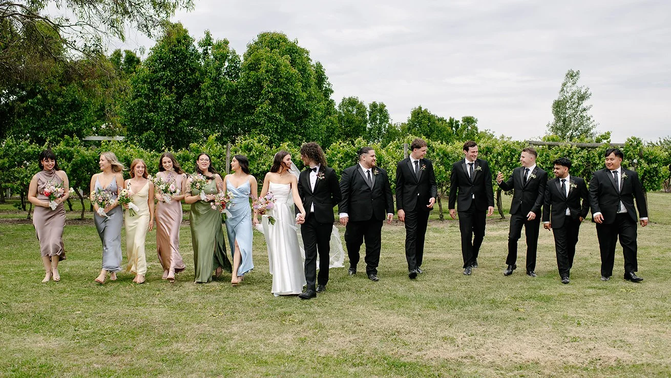 A wedding party walking outdoors on grass with trees in the background. The group includes women in colorful dresses holding flowers and men in black suits, some holding hands, all smiling and talking.